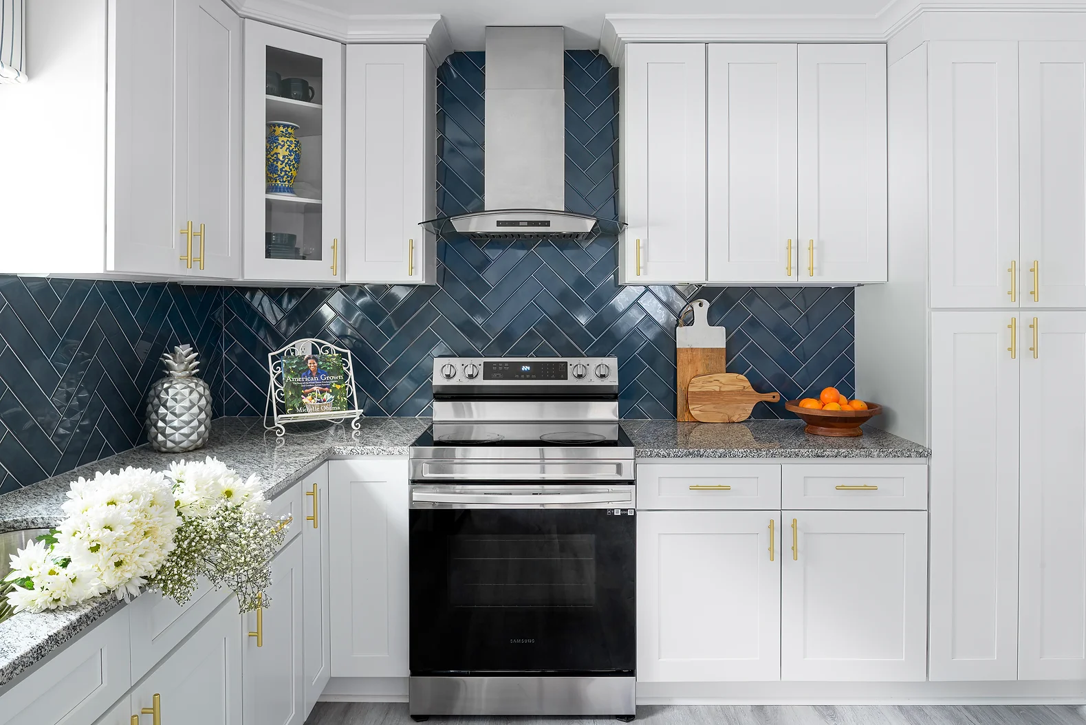 A modern kitchen featuring white cabinets, a stainless steel oven, and a blue herringbone backsplash. A dish of oranges and a decorative pineapple are on the counter, with white flowers in the foreground.