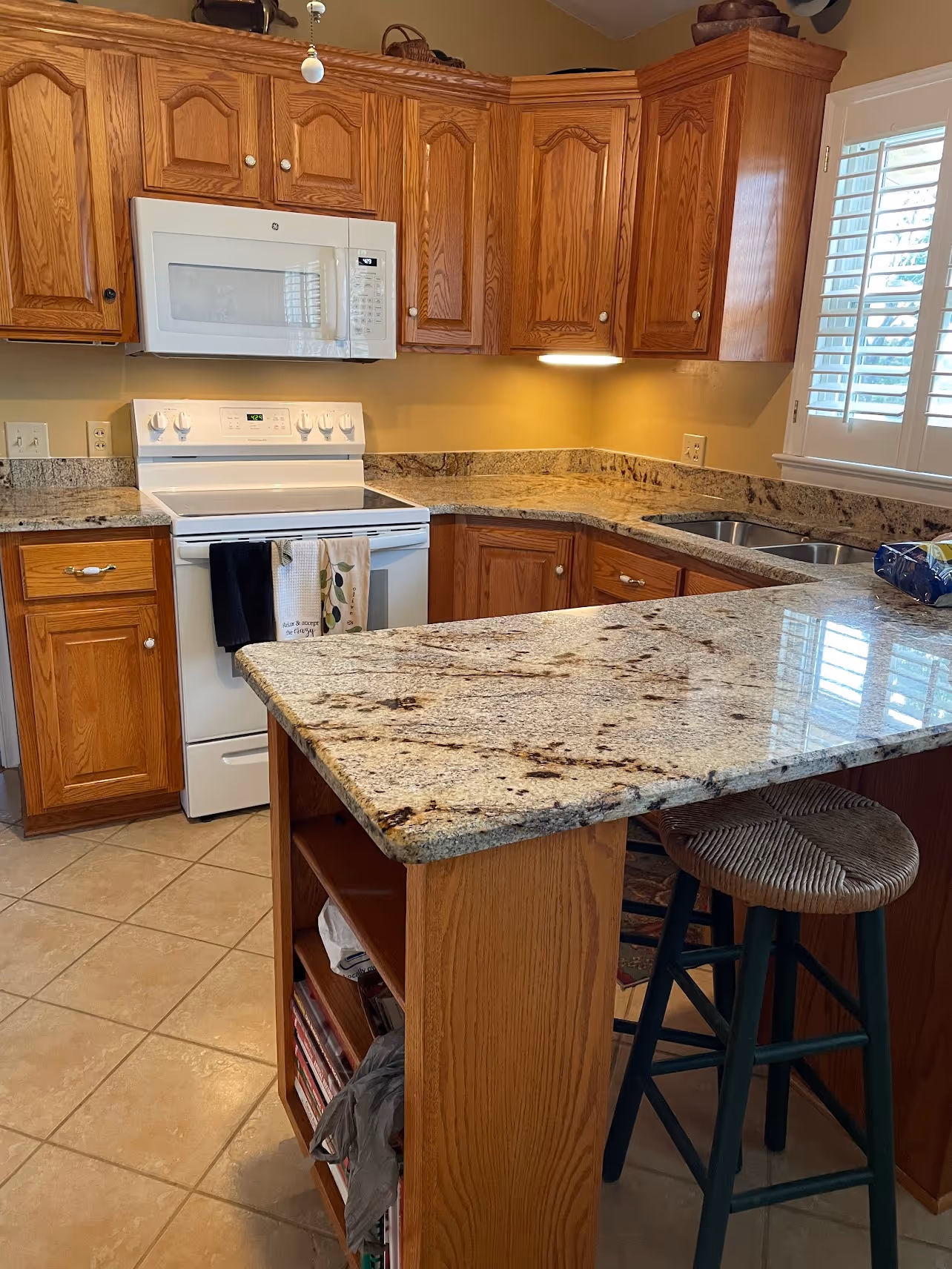 A corner view of a modern kitchen with wooden cabinets, granite countertops, a white stove, and a microwave. A bar stool is positioned at the countertop, and there are light-colored tiles on the floor.