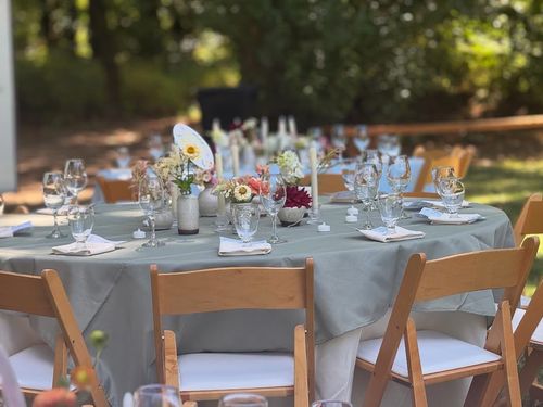 Outdoor round table set with a gray tablecloth, wooden chairs, glassware, napkins, candles, and floral centerpieces.