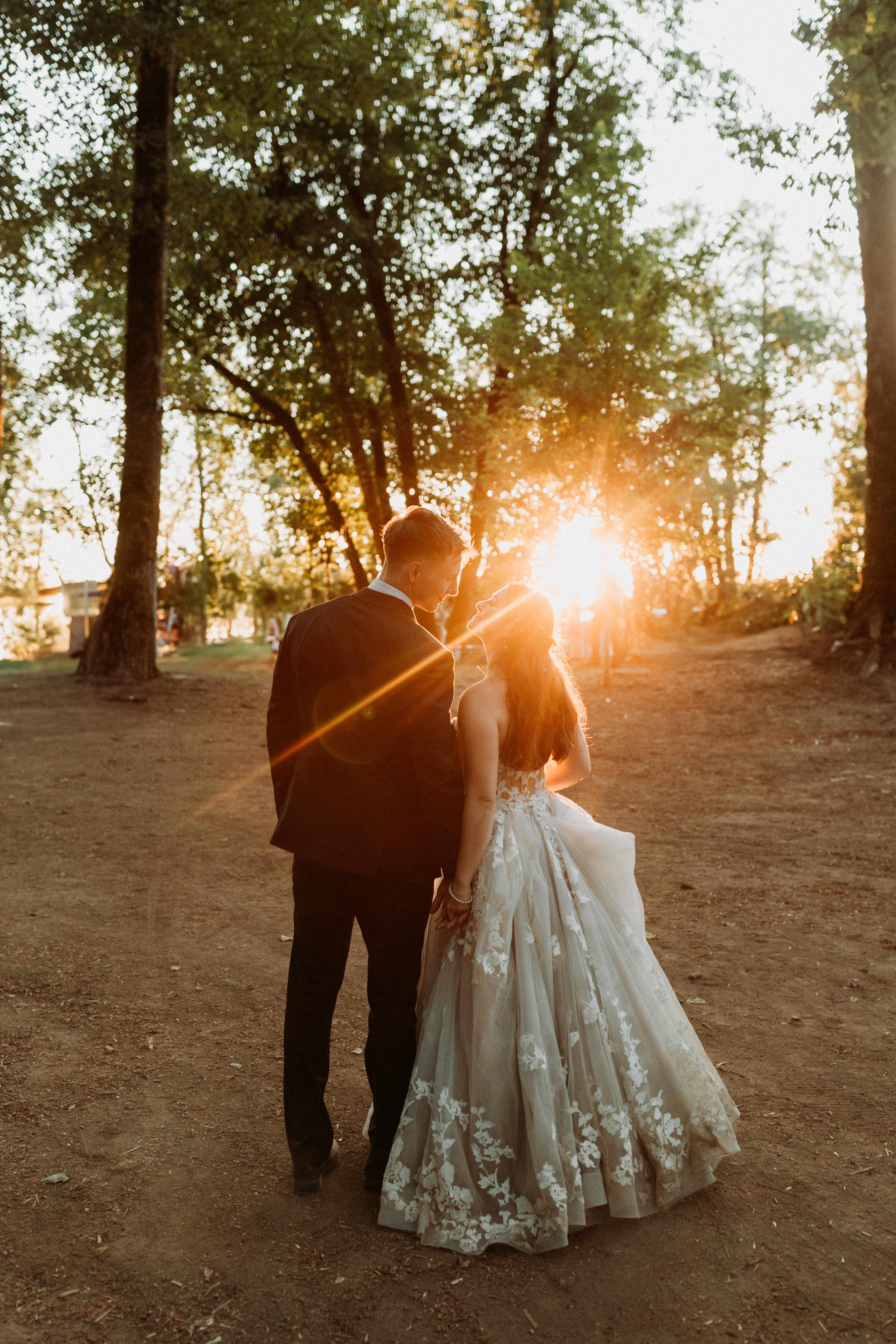 Couple in wedding attire holding hands and gazing at each other in a sunlit forest setting during sunset.