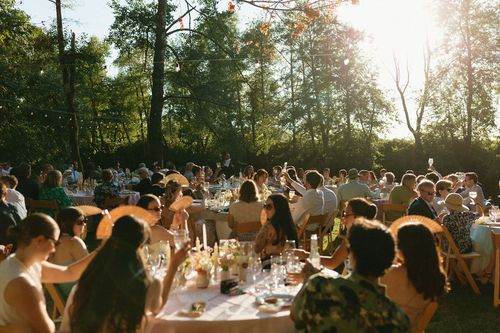 People seated at outdoor tables in a sunlit garden, raising glasses in a toast during a daytime celebration.