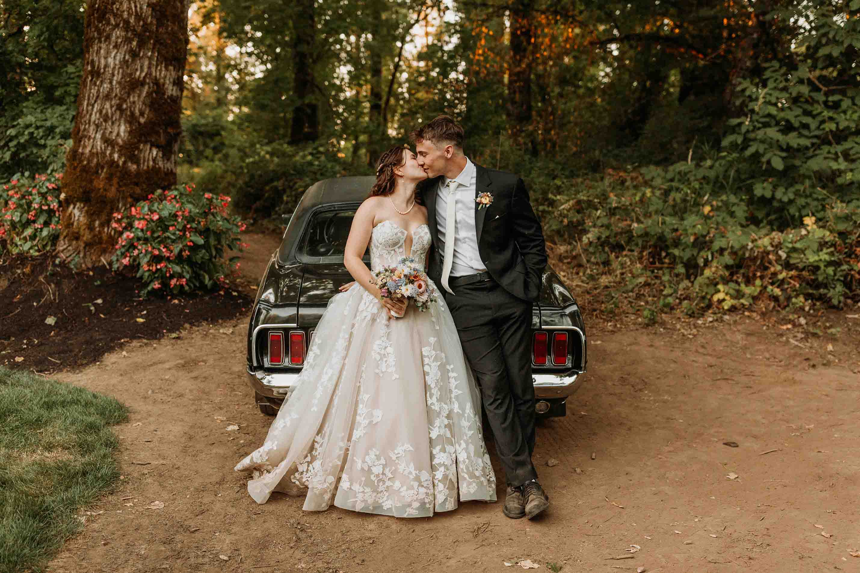 Bride in floral lace wedding dress and groom in black suit kissing while leaning against a vintage car in a wooded area.