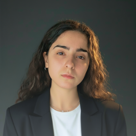 Portrait of a young woman with long brown hair wearing a dark blazer and white top against a dark background.