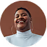 Smiling person with short hair wearing a white turtleneck against a brown background.