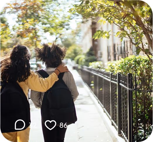 Two children with backpacks walking arm-in-arm on a sunny sidewalk lined with trees and a wrought-iron fence.