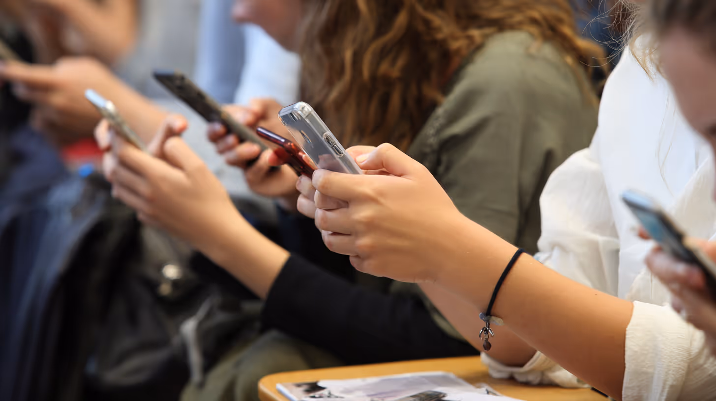 Gros plan sur des mains d’élèves utilisant leurs smartphones en classe, représentant la tentation constante et la fragmentation de l’attention que les établissements cherchent à réguler.