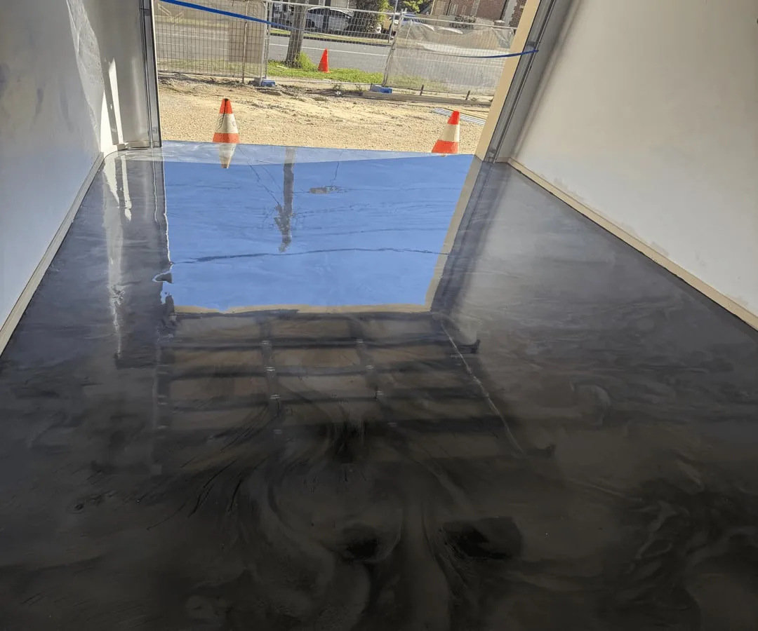 Gunmetal metallic epoxy floor reflecting the open garage doorway and daylight, with traffic cones visible outside.