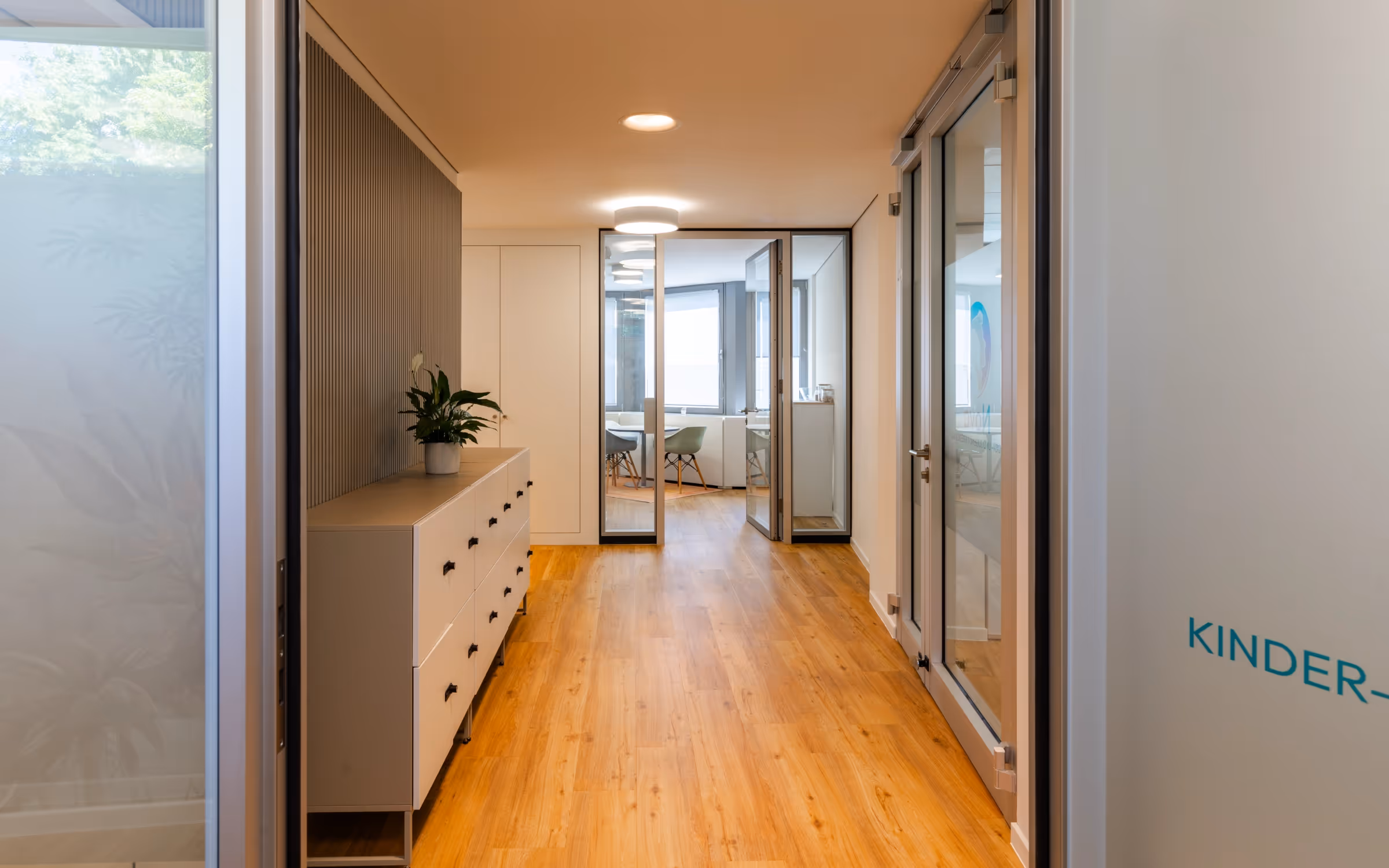 Heller Praxisflur mit Holzboden, weißer Kommode und Blick in einen Therapieraum.
