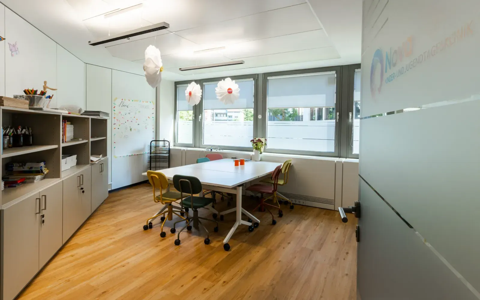 Heller Gruppenraum mit Tisch, bunten Stühlen und einem Whiteboard für kreative Aktivitäten.