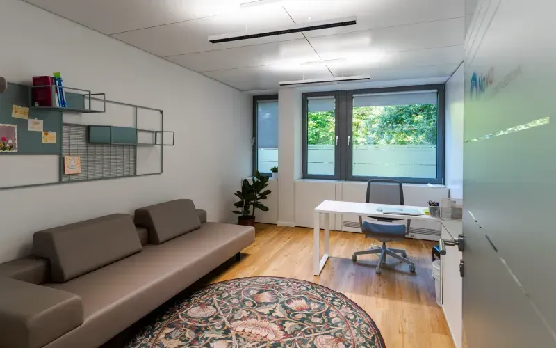 Therapieraum mit Sofa, Schreibtisch und einem großen, floralen Rundteppich.
