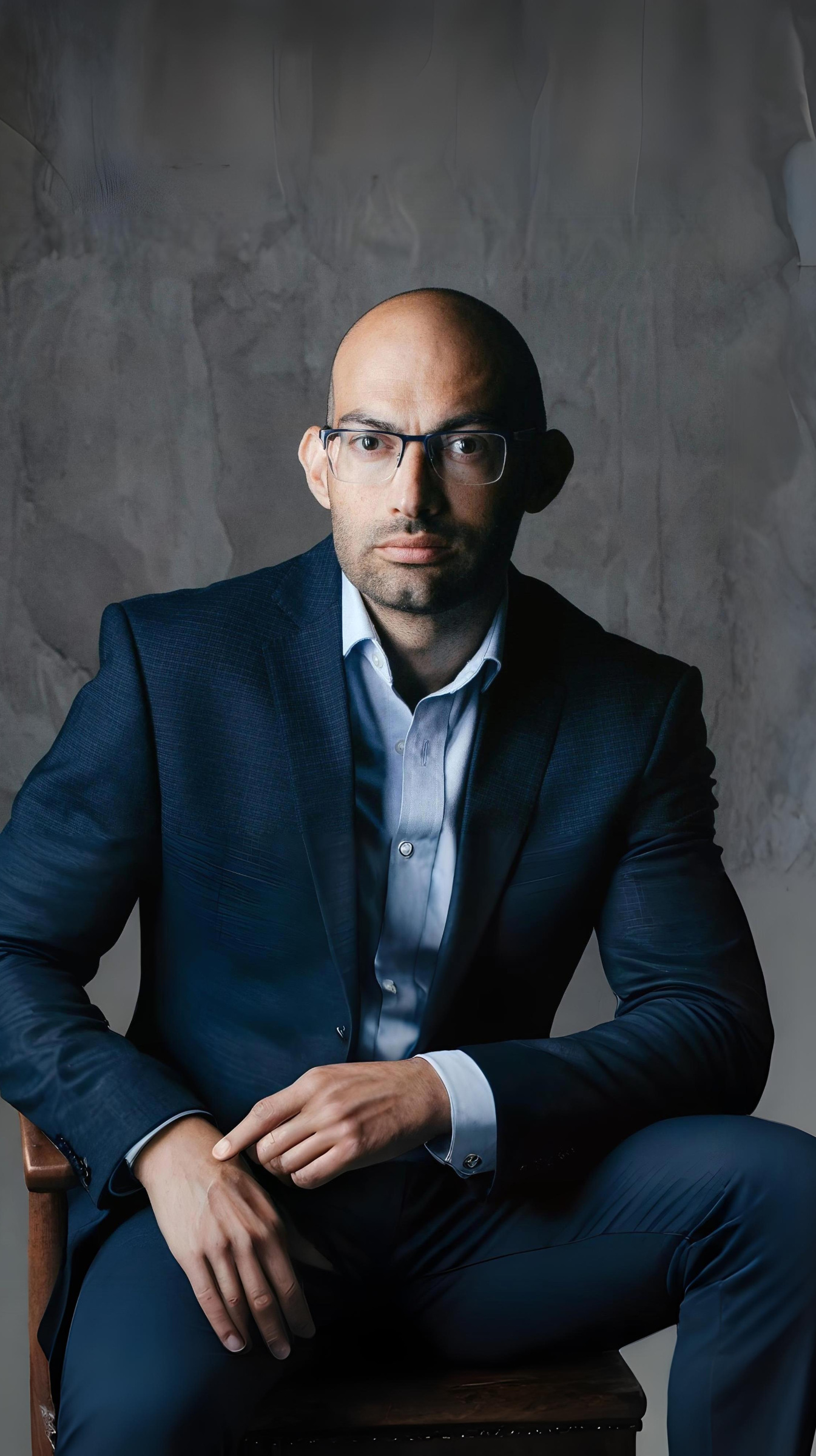 Bald man with glasses in a dark blue suit seated on a chair against a textured gray background.