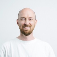 Smiling bald man with trimmed beard wearing a white shirt against a plain light background.