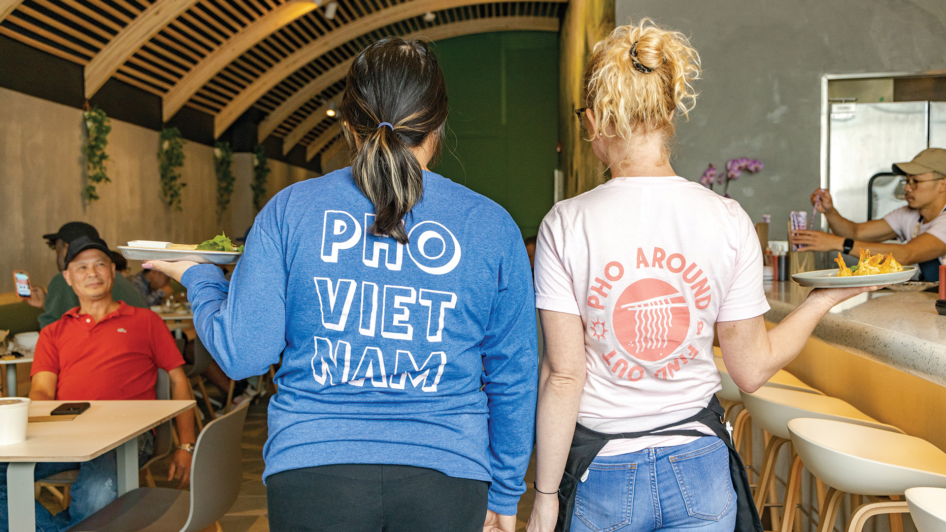 Two restaurant staff members carrying food, seen from behind, wearing shirts with "Pho Vietnam" and "Pho Around, Find Out" slogans. Diners are seated at tables in the background.