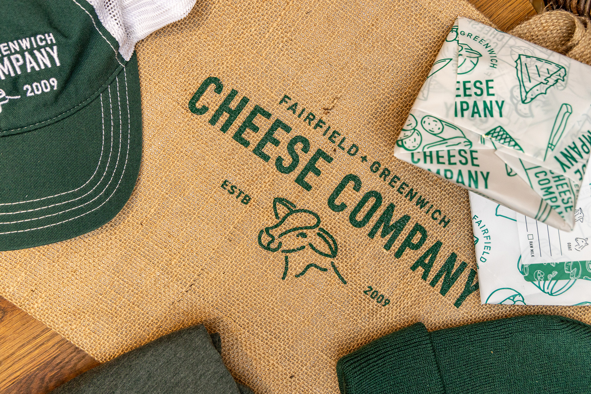 Flat lay of Fairfield Cheese Co. branded items, including a green and white trucker hat, a burlap tote bag with the logo, and cheese packaging with a cow illustration, showcasing the company's visual identity.