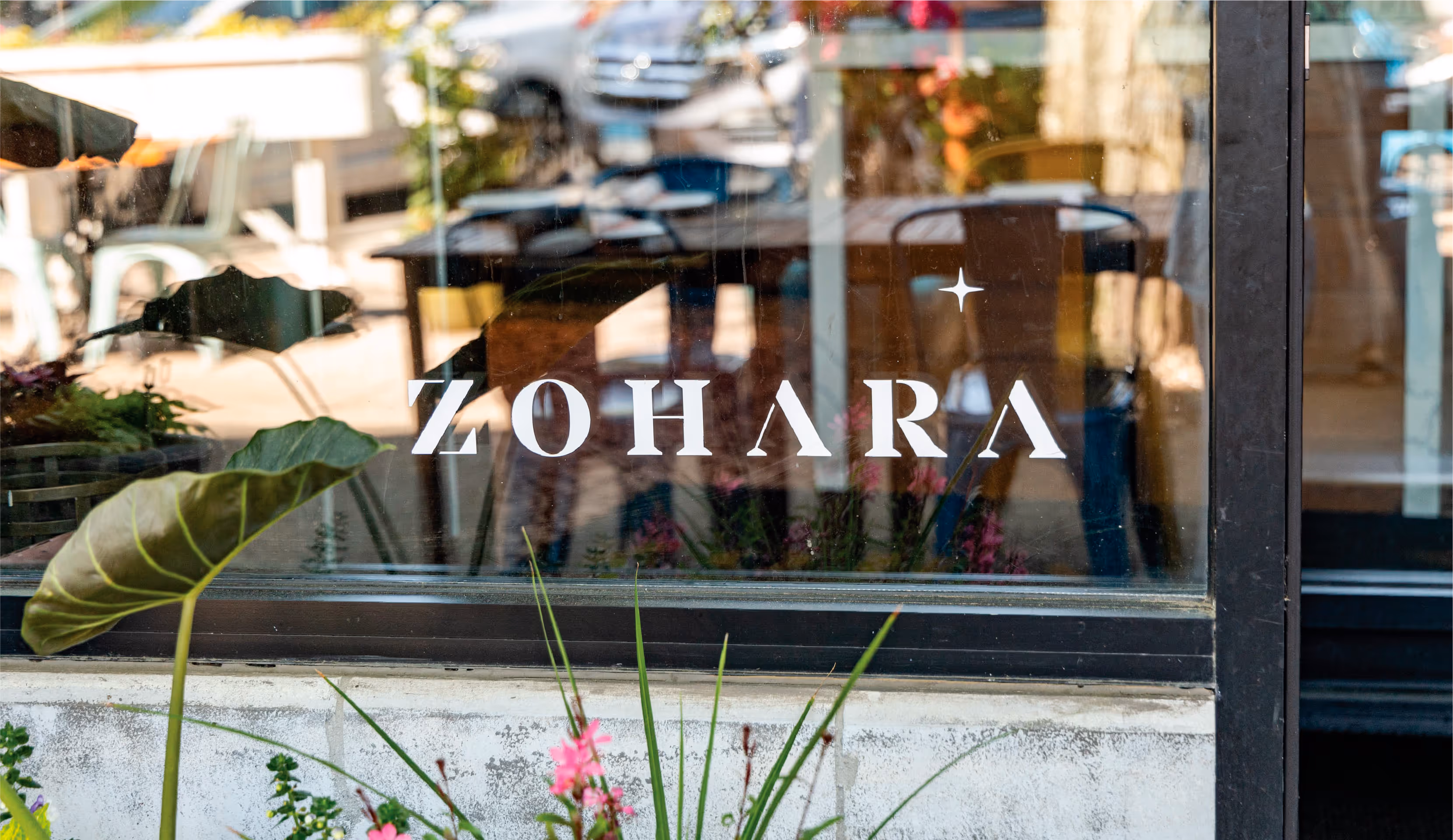 A view of the Zohara storefront through a large glass window. The restaurant's name, 'ZOHARA,' is prominently displayed in white lettering on the glass, with a small star above the 'A'.