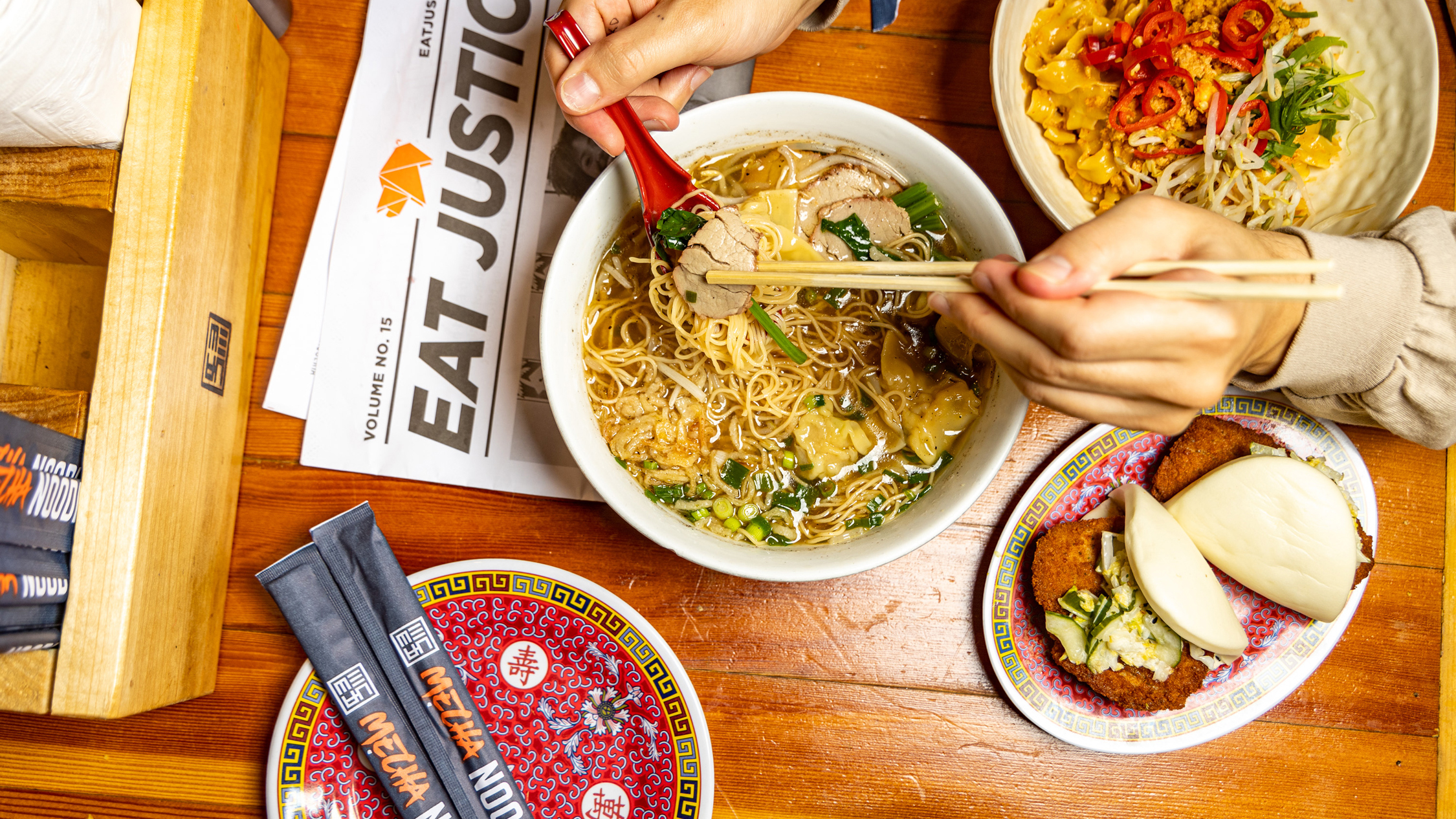 Stylized overhead shot of a culinary spread from Mecha Noodle Bar, showcasing their signature ramen, flavorful side dishes, and branded newspaper with the headline "EAT JUSTICE," all arranged on a rustic wooden table.