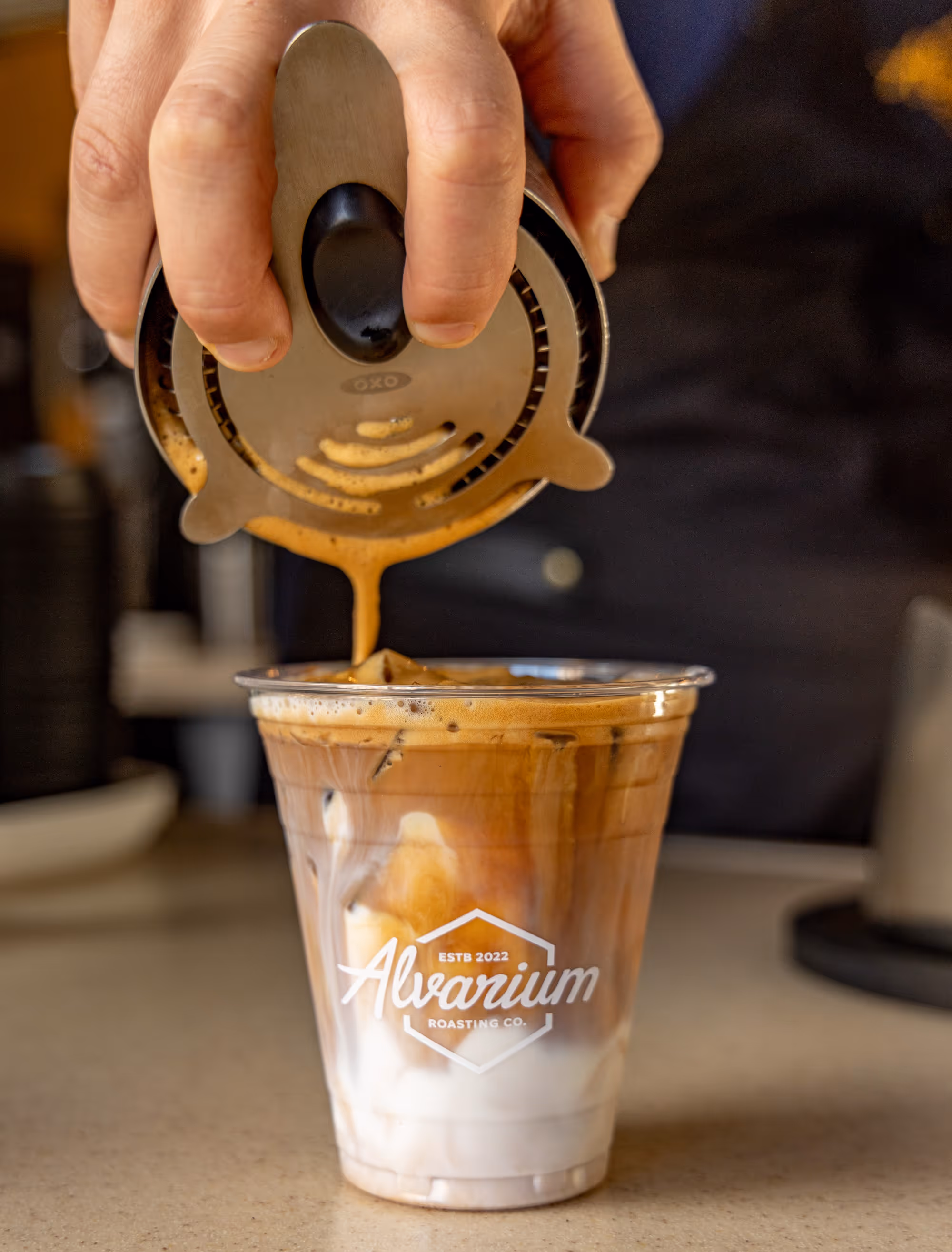 Photo of a beverage preparation, emphasizing the combination of the pour-over coffee technique, the refreshing iced coffee drink with its distinct layers, and the Alvarium Roasting Co. branding on the cup.