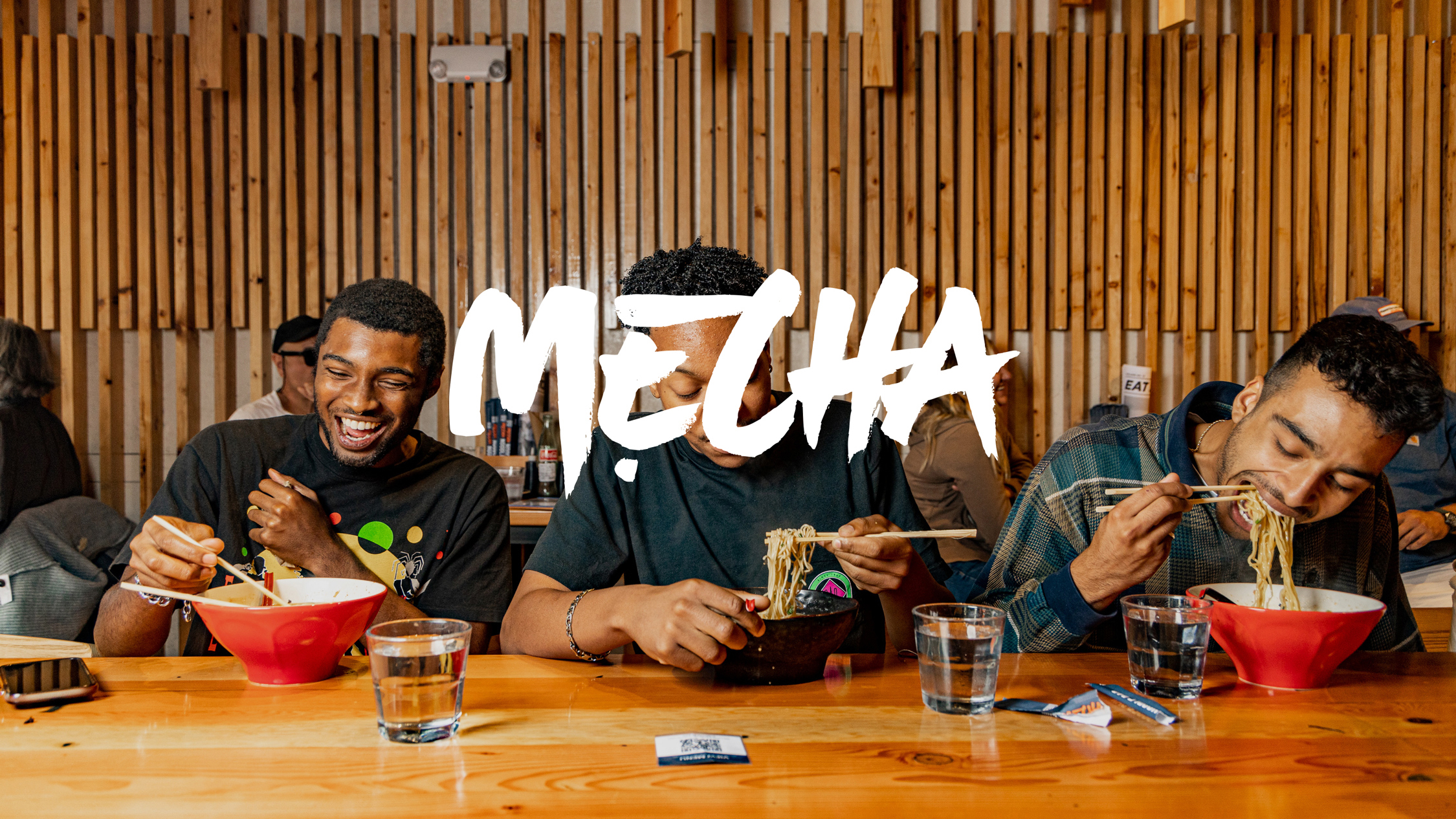 Customers eating ramen at a wooden table with red and black bowls, glasses of water, and with the word "MECHA" overlaid.