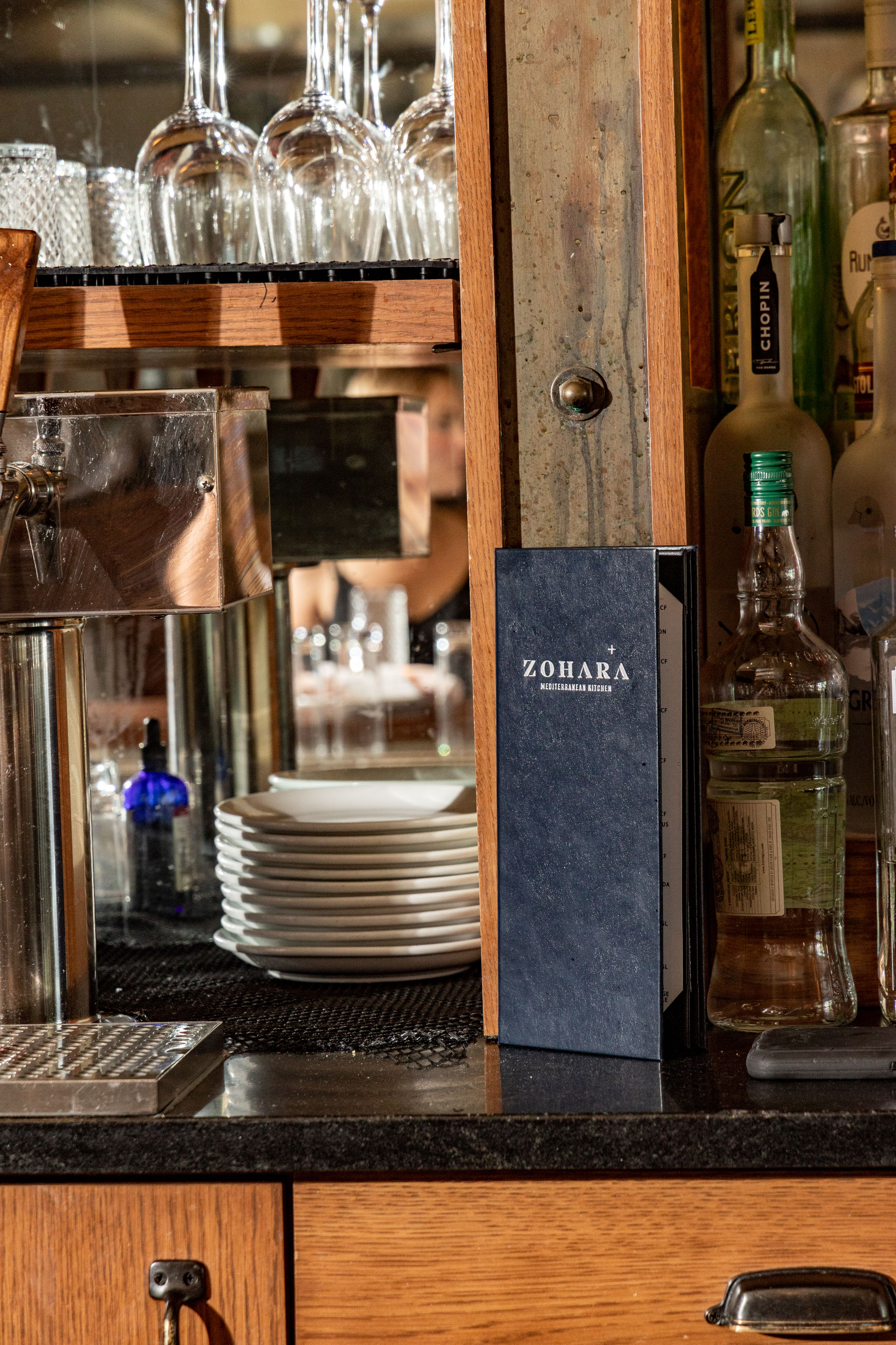 A dark blue Zohara menu stands upright on a bar counter, surrounded by glassware, bottles of alcohol, and other bar equipment.