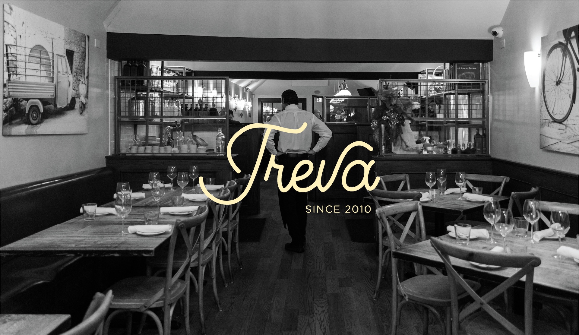 Black and white photograph of Treva restaurant's dining area, showcasing tables with place settings, bentwood chairs, and a glimpse into the kitchen, overlaid with the Treva logo in a script font.