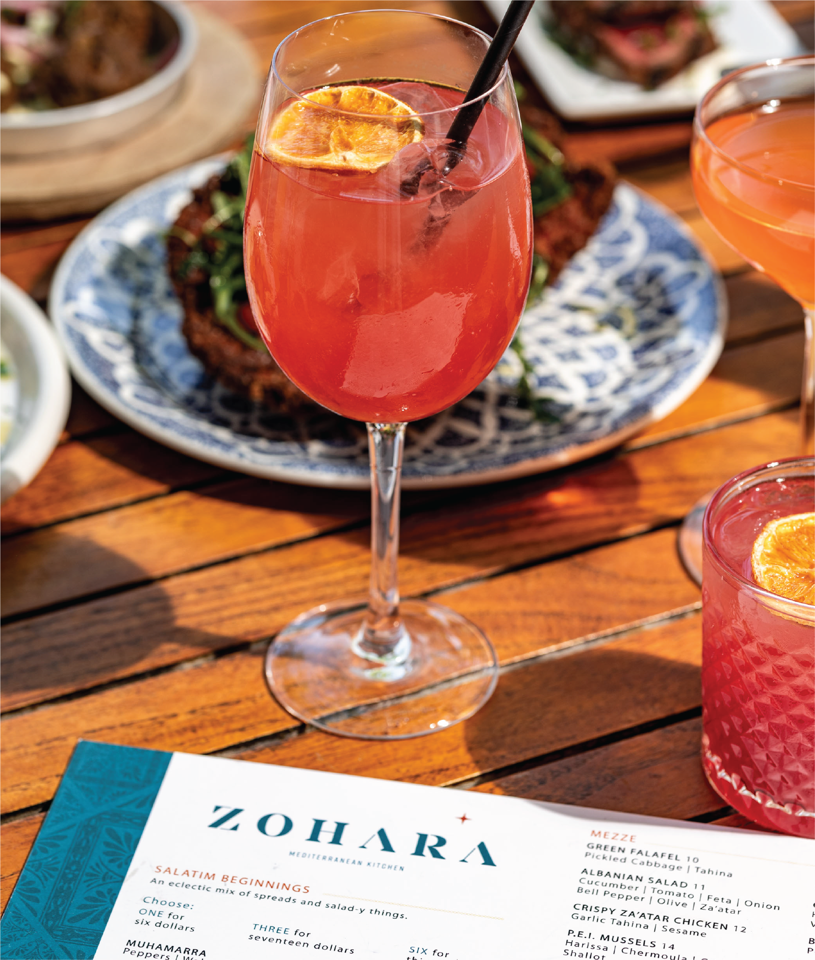 A close-up shot of a tall glass filled with a vibrant pink cocktail. The drink is garnished with a lemon slice and a straw. In the background, you can see the Zohara menu and plates of food.
