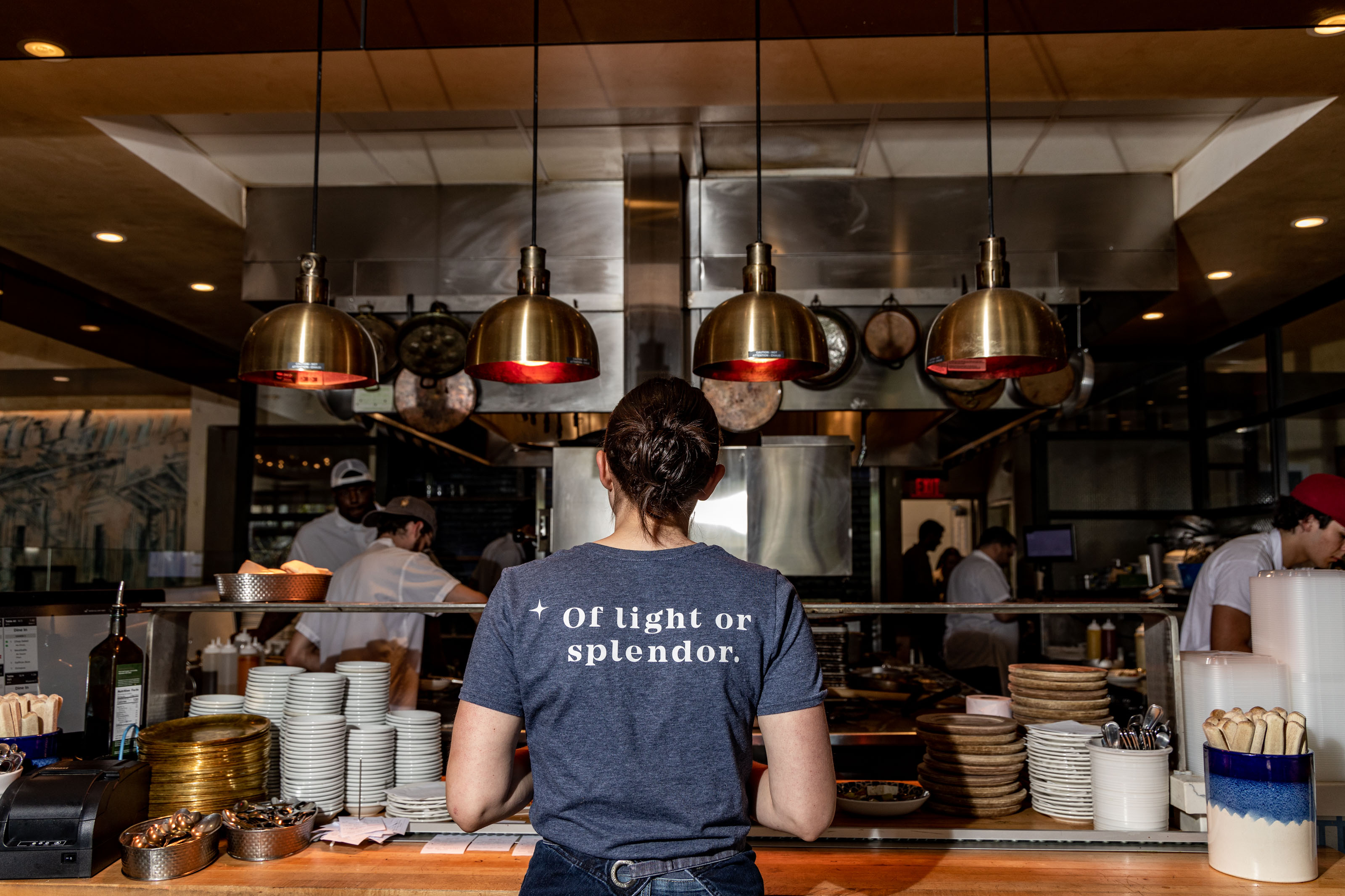 A view into a restaurant's open kitchen, showing staff working and preparing food. Copper pendant lights hang above a counter where a person wearing a t-shirt with the phrase 'Of light or splendor' stands with their back to the viewer.
