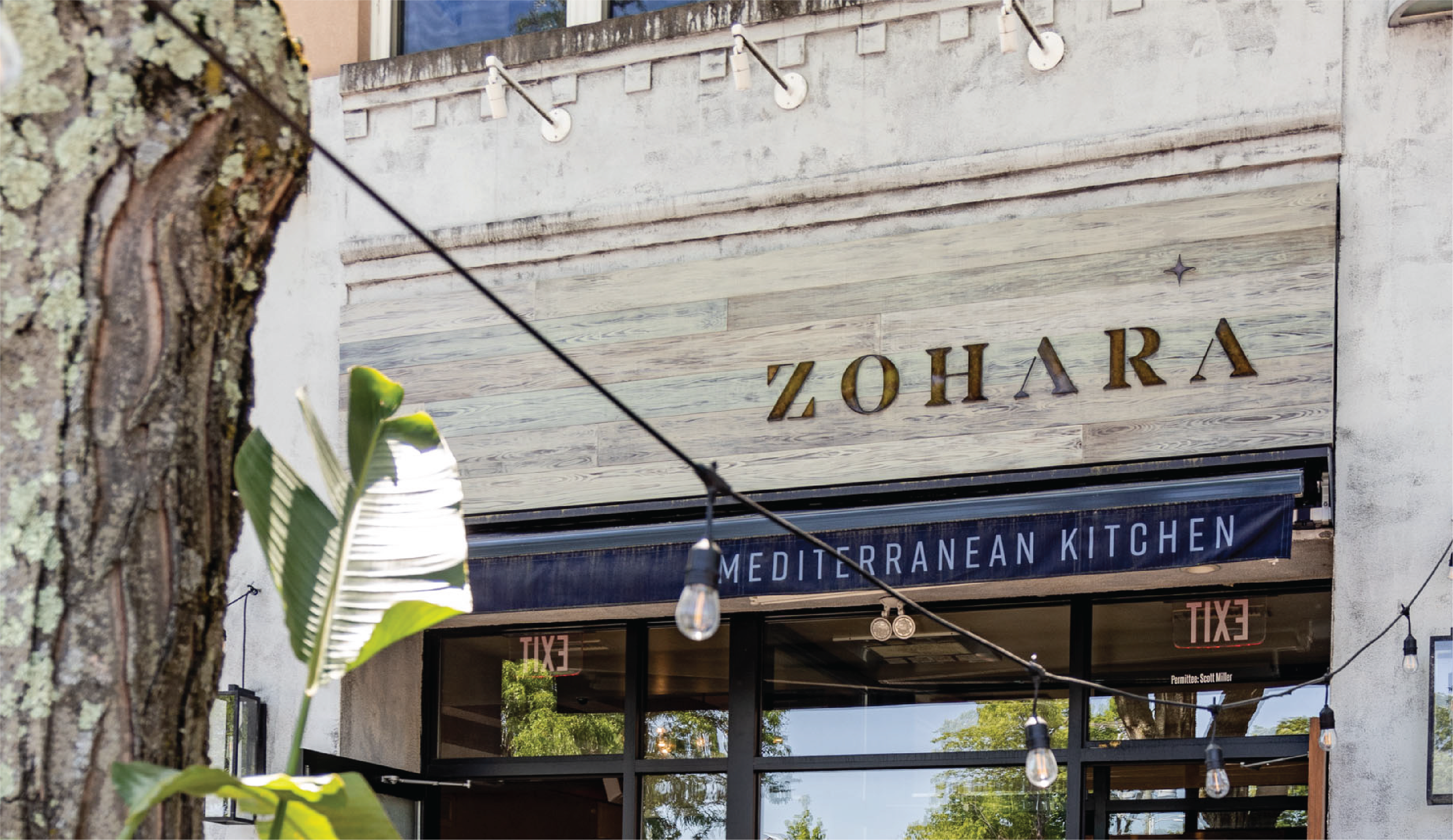 The exterior of Zohara Mediterranean Kitchen is captured in a composition that highlights the interplay between the building's architectural elements and the restaurant's signage. The textured concrete facade, combined with the wooden sign and dark awning, creates a visually interesting and inviting entrance. String lights add a touch of ambiance.