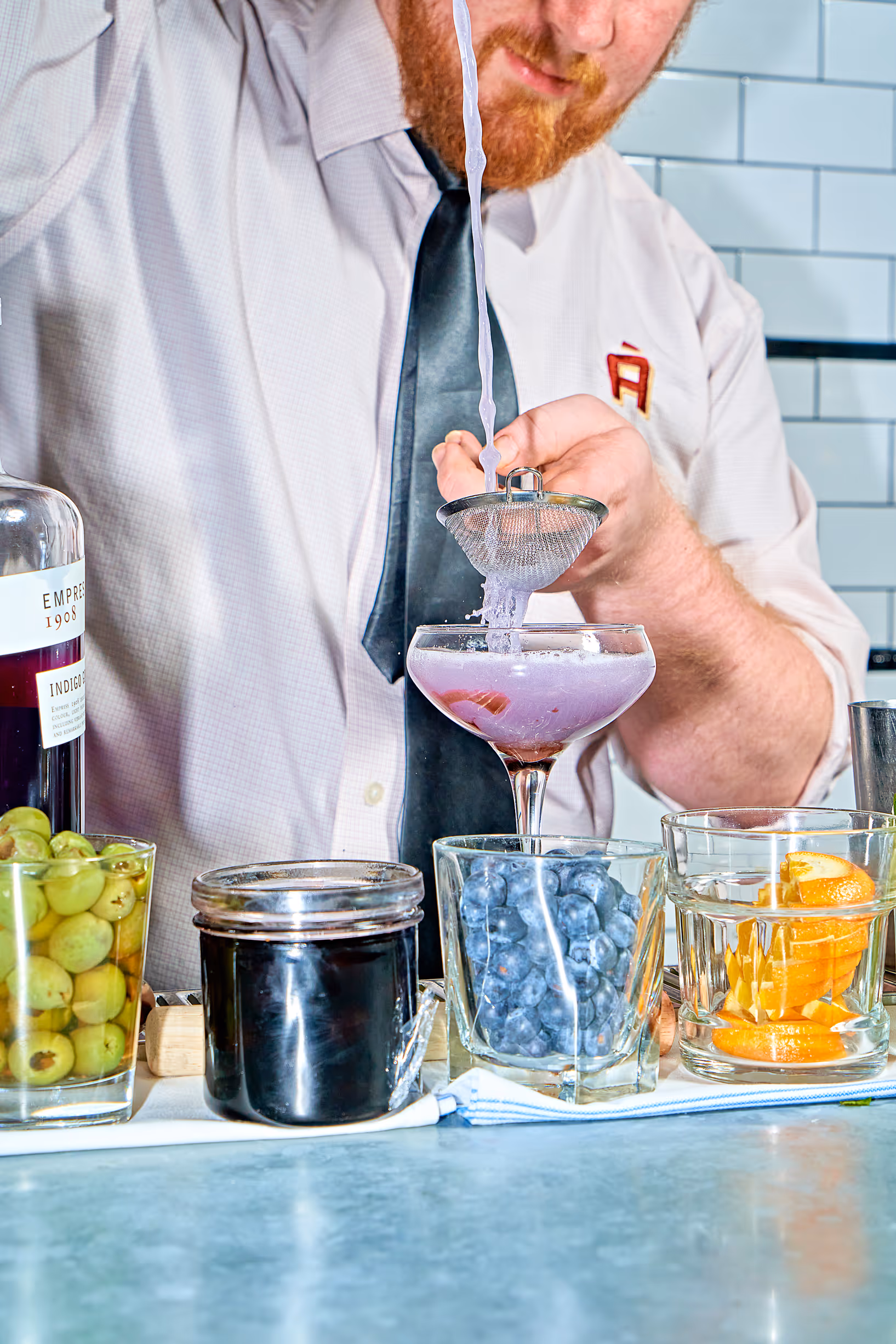 A close-up photograph capturing the moment a bartender meticulously strains a vibrant purple cocktail from a shaker into a martini glass. The scene is filled with colorful ingredients, creating a sense of lively energy.