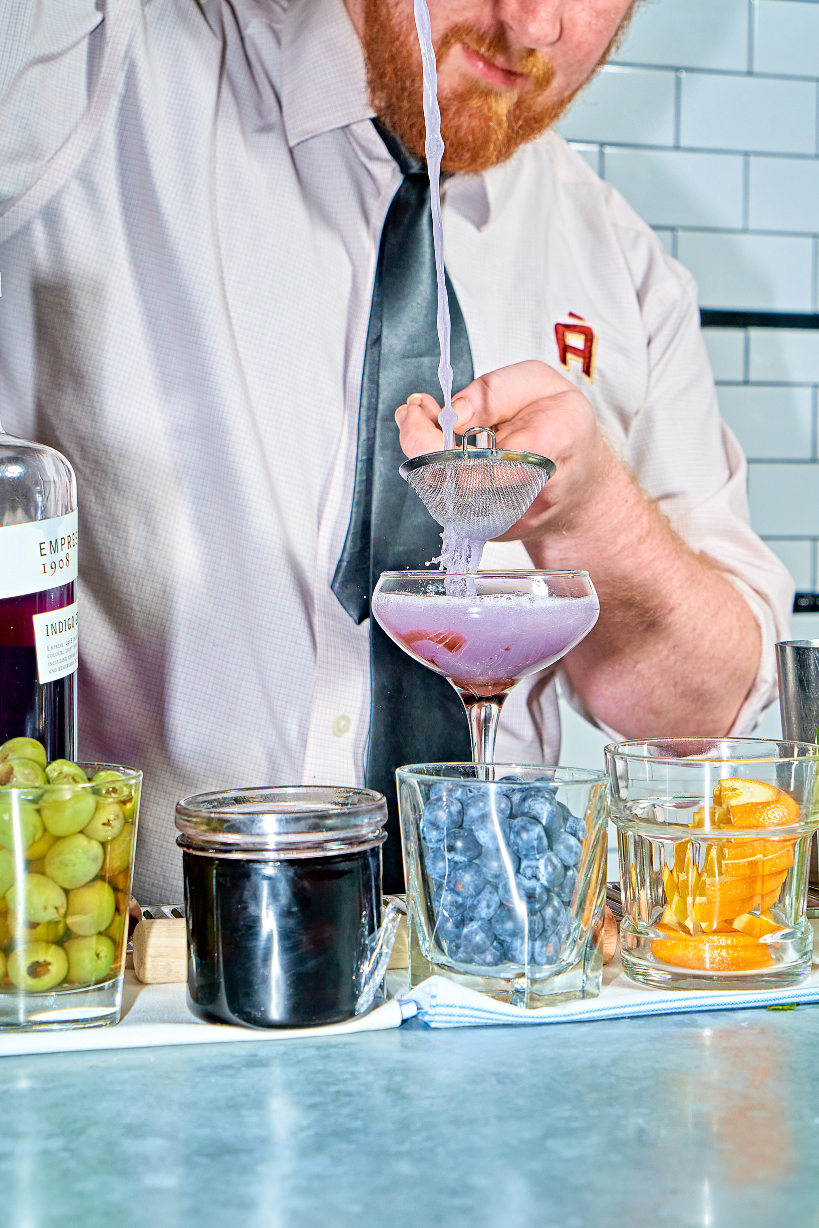 A close-up photograph capturing the moment a bartender meticulously strains a vibrant purple cocktail from a shaker into a martini glass. The scene is filled with colorful ingredients, creating a sense of lively energy.