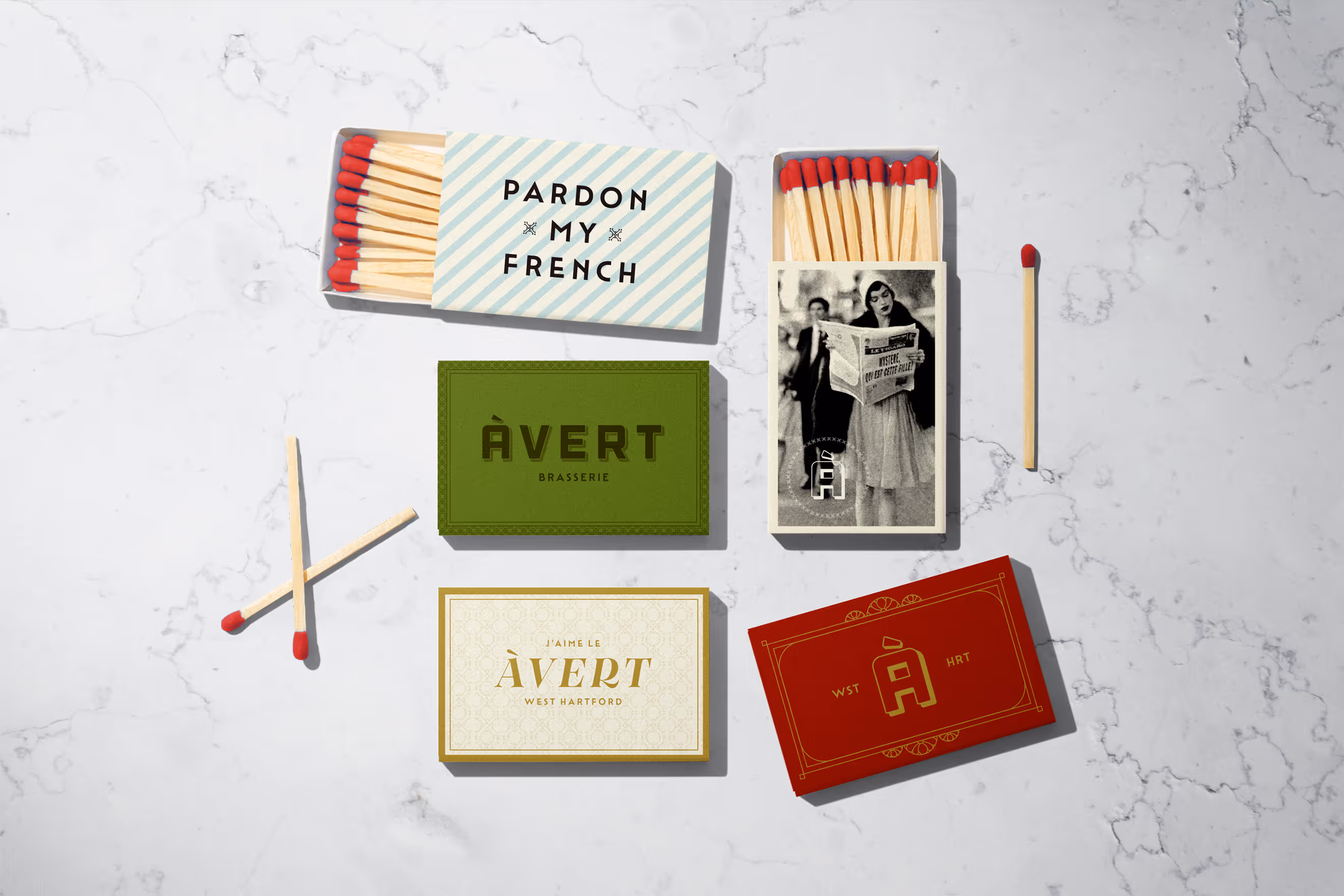 A digitally created graphic of five matchbox covers on a white marble surface. The matchboxes showcase a cohesive brand identity with a vintage aesthetic. Each box features distinct design elements, including a woman reading a newspaper, a bold "Avert" logo, and a playful "Pardon My French" text.