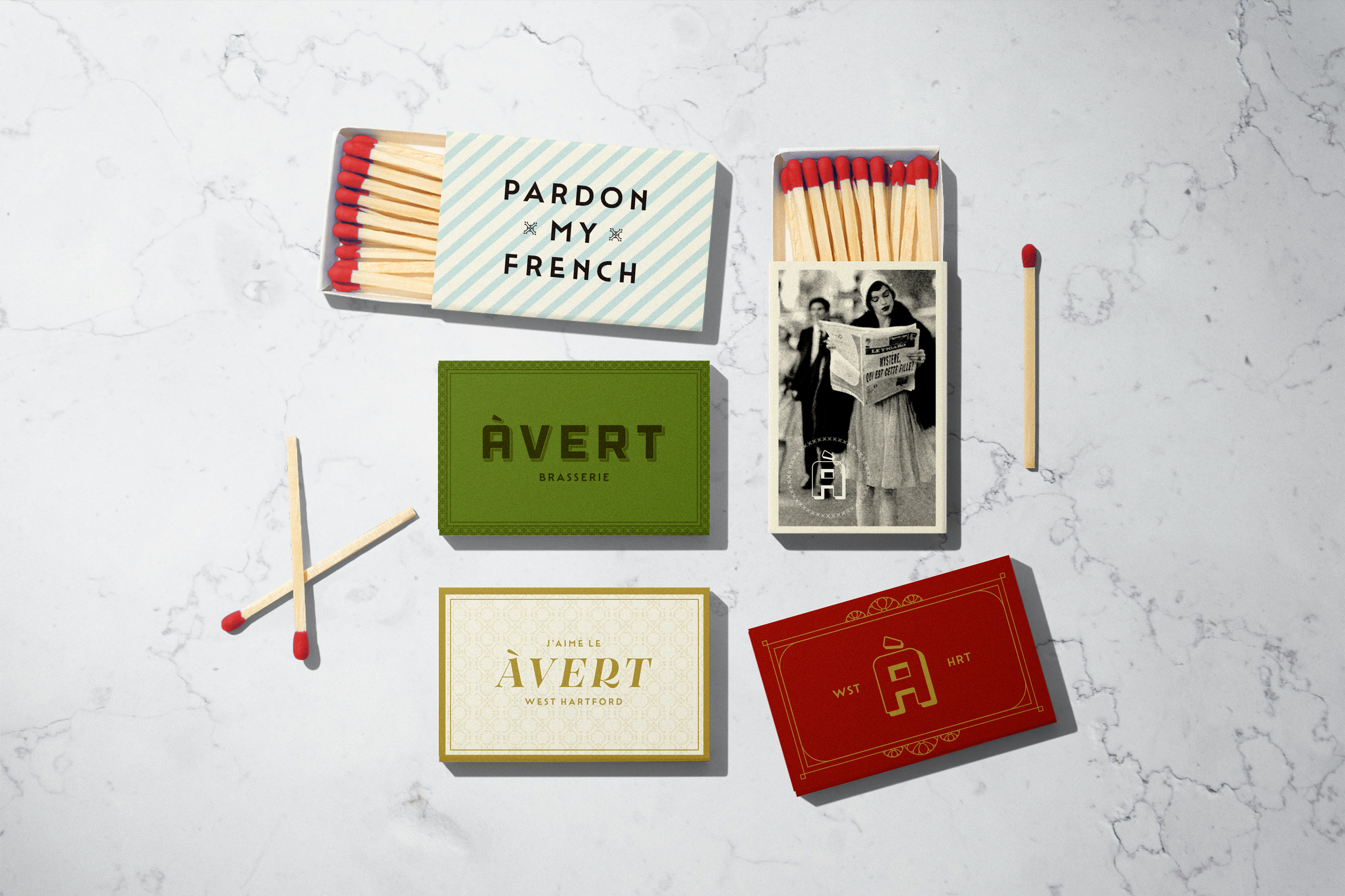 A digitally created graphic of five matchbox covers on a white marble surface. The matchboxes showcase a cohesive brand identity with a vintage aesthetic. Each box features distinct design elements, including a woman reading a newspaper, a bold "Avert" logo, and a playful "Pardon My French" text.
