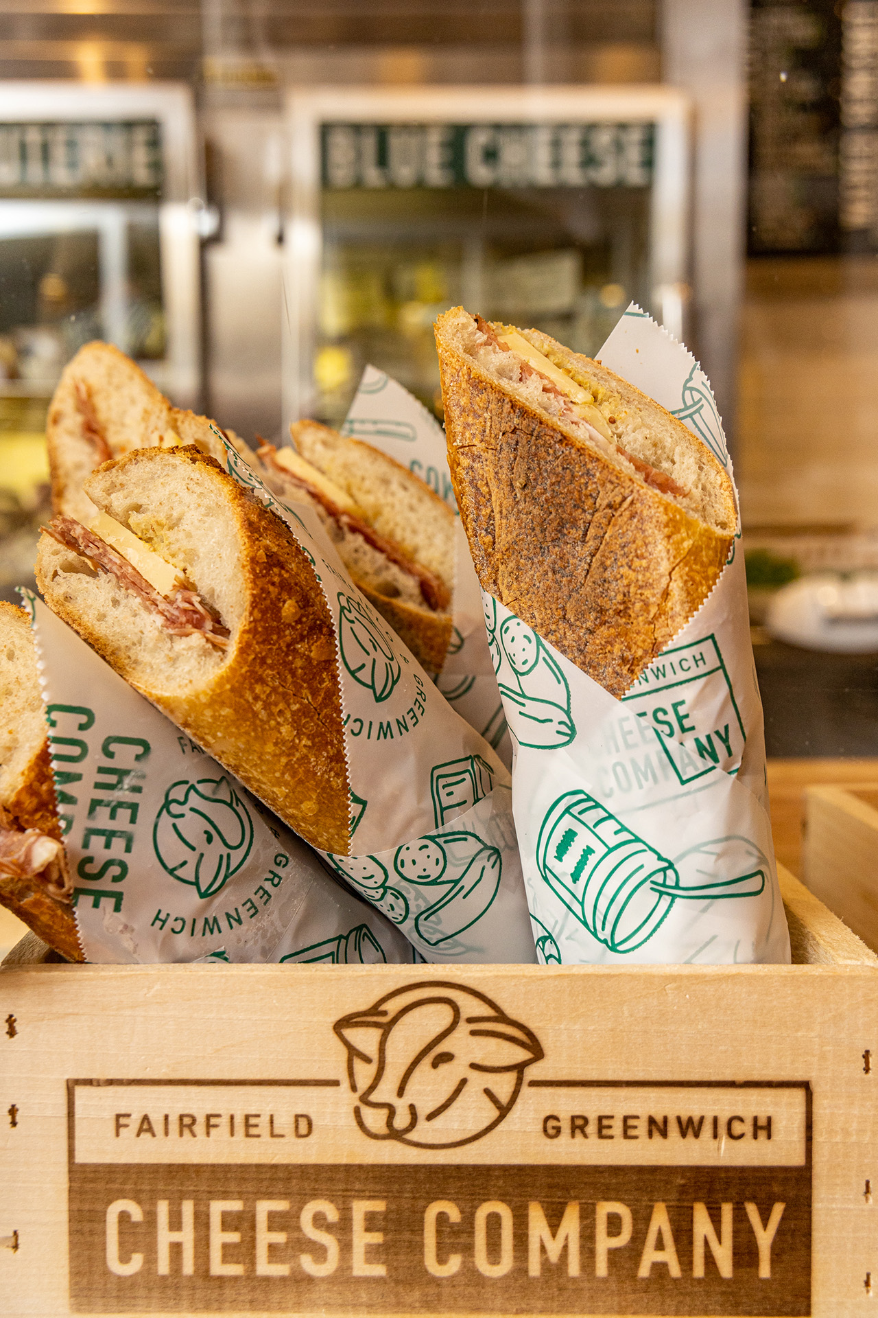 Close-up of several sandwiches wrapped in branded Fairfield Cheese Company paper, showcasing the sandwiches' fillings and textures alongside the company's logo.