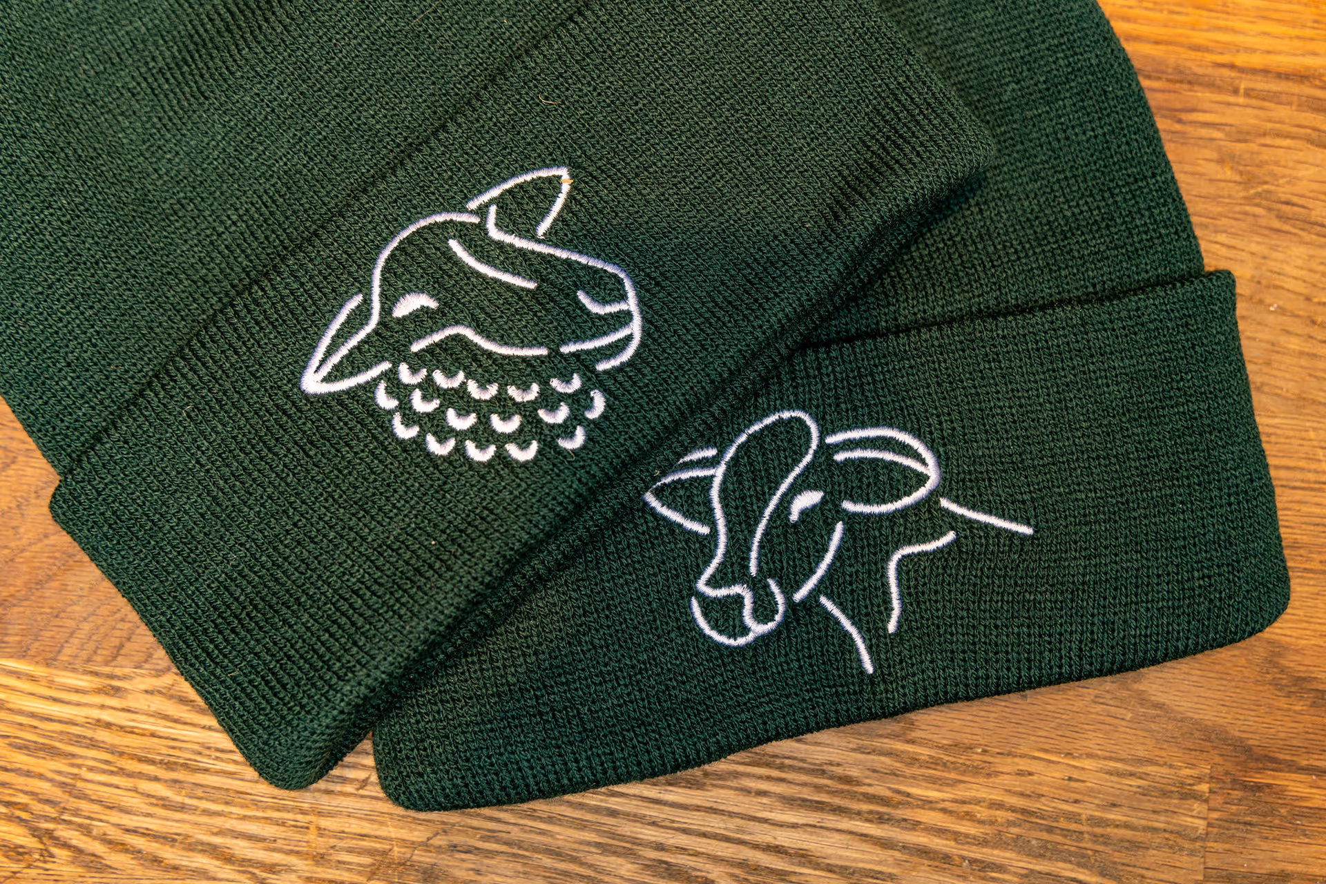 Close-up of two dark green knit beanies, one featuring an embroidered sheep design, the other an embroidered cow head design, both in white thread.