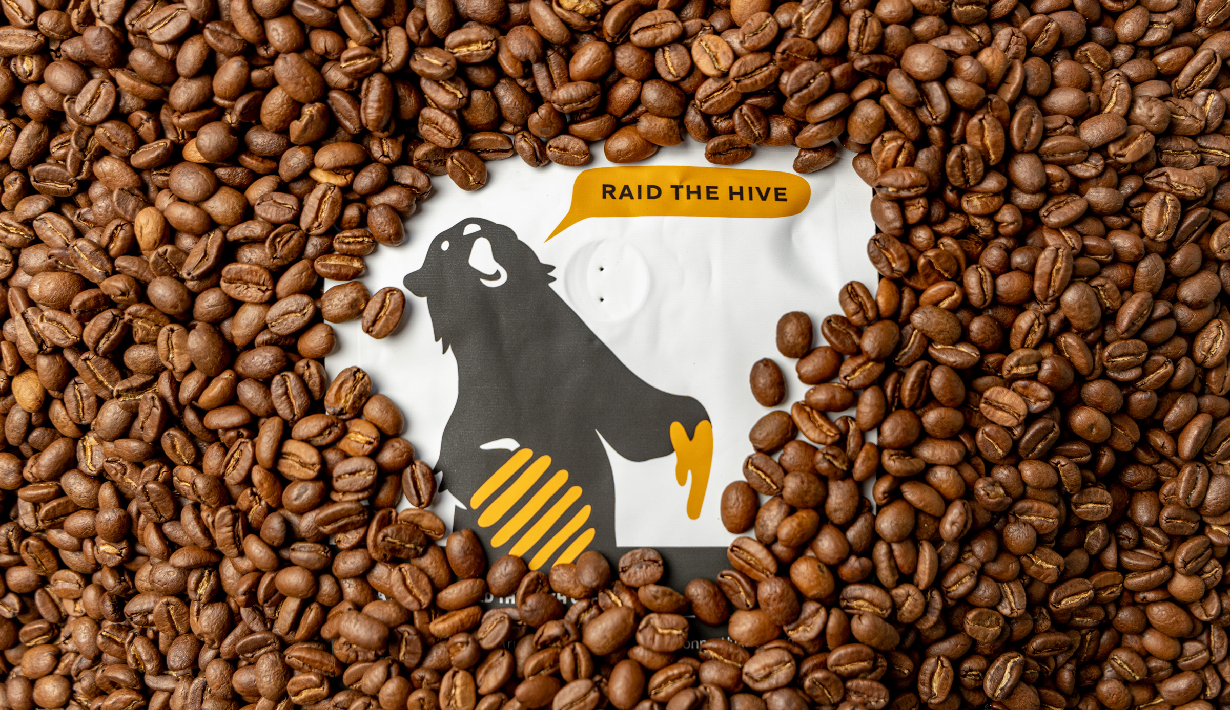 Image of an Alvarium Roasting Co. coffee bag nestled amongst roasted coffee beans, showcasing the brand's bear logo and "RAID THE HIVE" slogan.
