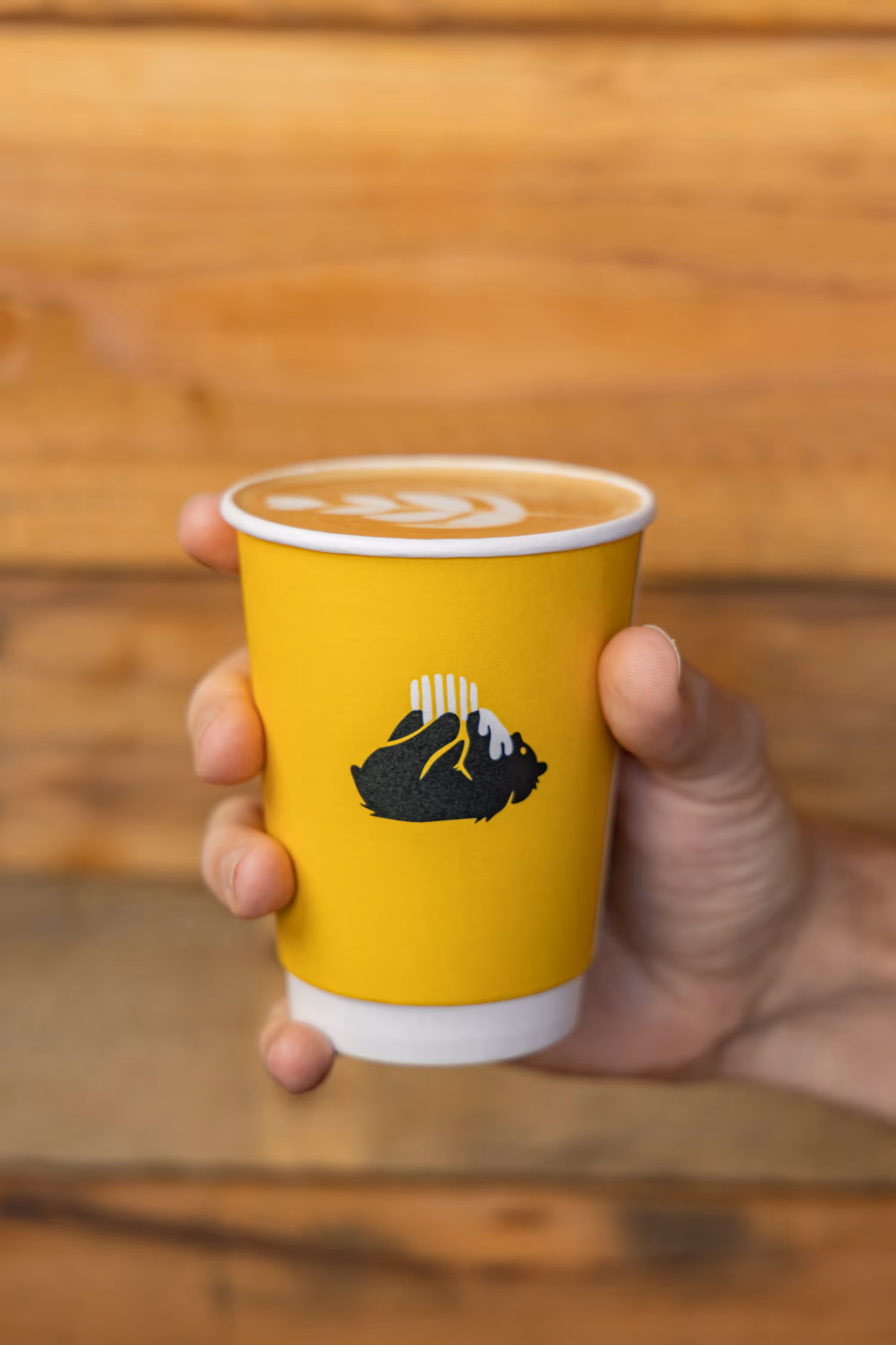 Image of a hand holding a yellow paper coffee cup featuring the Alvarium Roasting Co. bear logo, showcasing the brand's visual identity and the cup's design against a blurred wooden background.