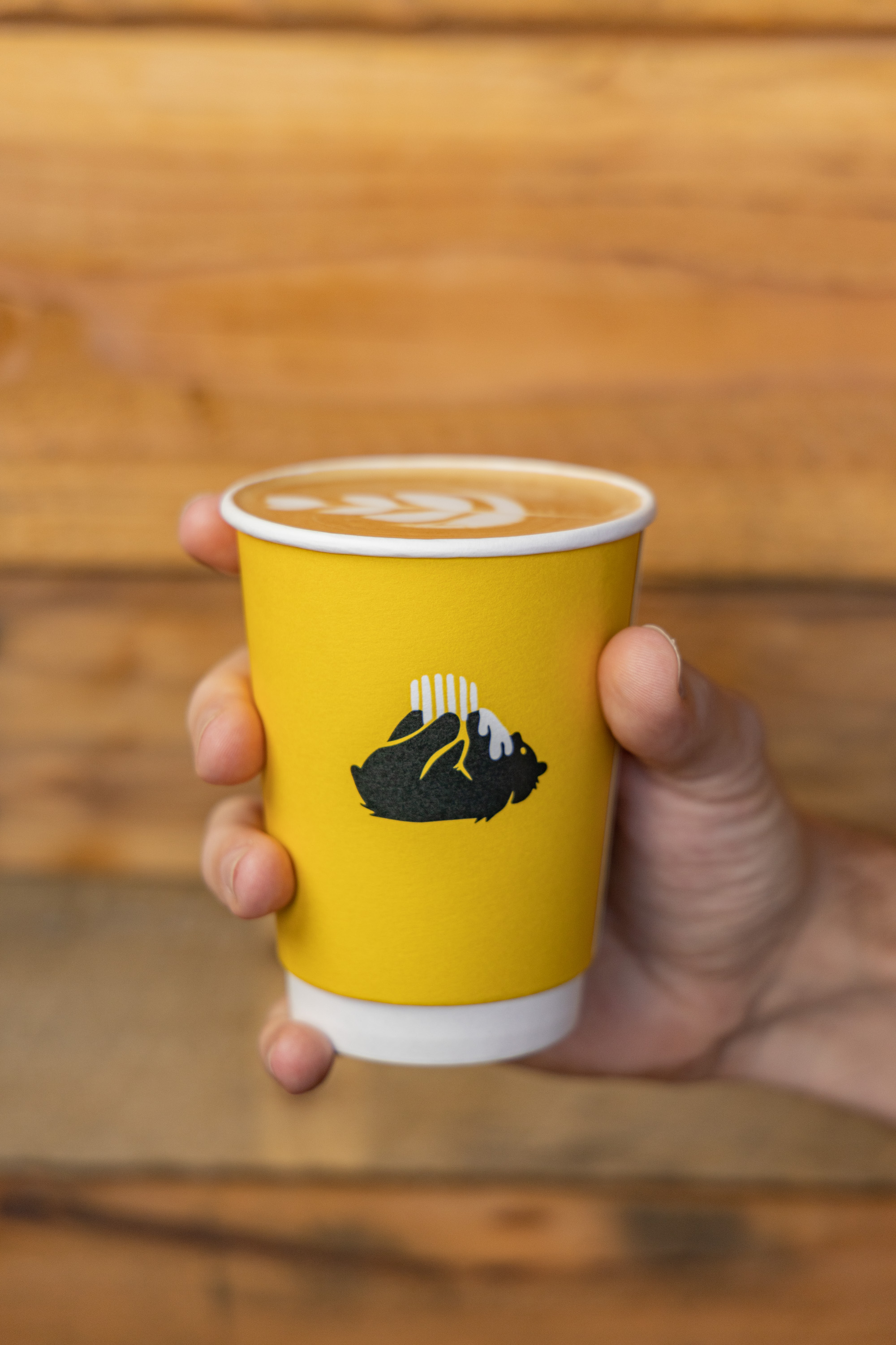 Image of a hand holding a yellow paper coffee cup featuring the Alvarium Roasting Co. bear logo, showcasing the brand's visual identity and the cup's design against a blurred wooden background.