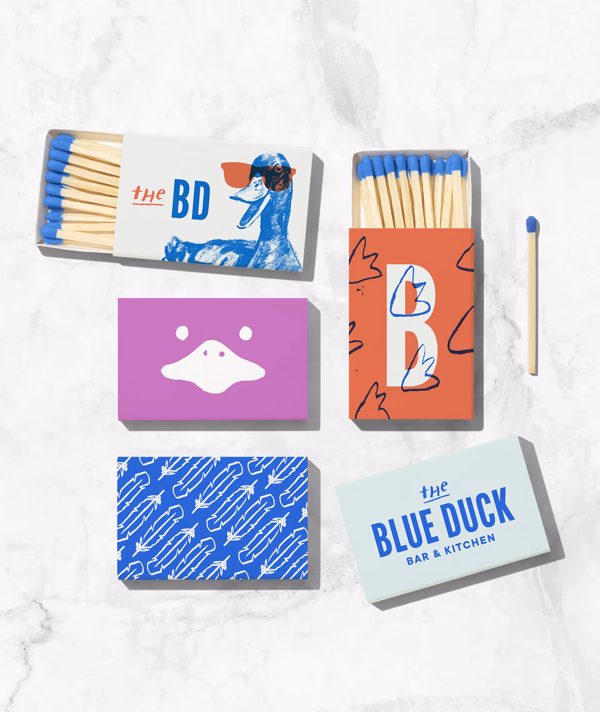 Photo of multiple matchboxes featuring The Blue Duck branding through typography, illustration, and color accents, demonstrating the brand's playful and recognizable aesthetic on packaging.