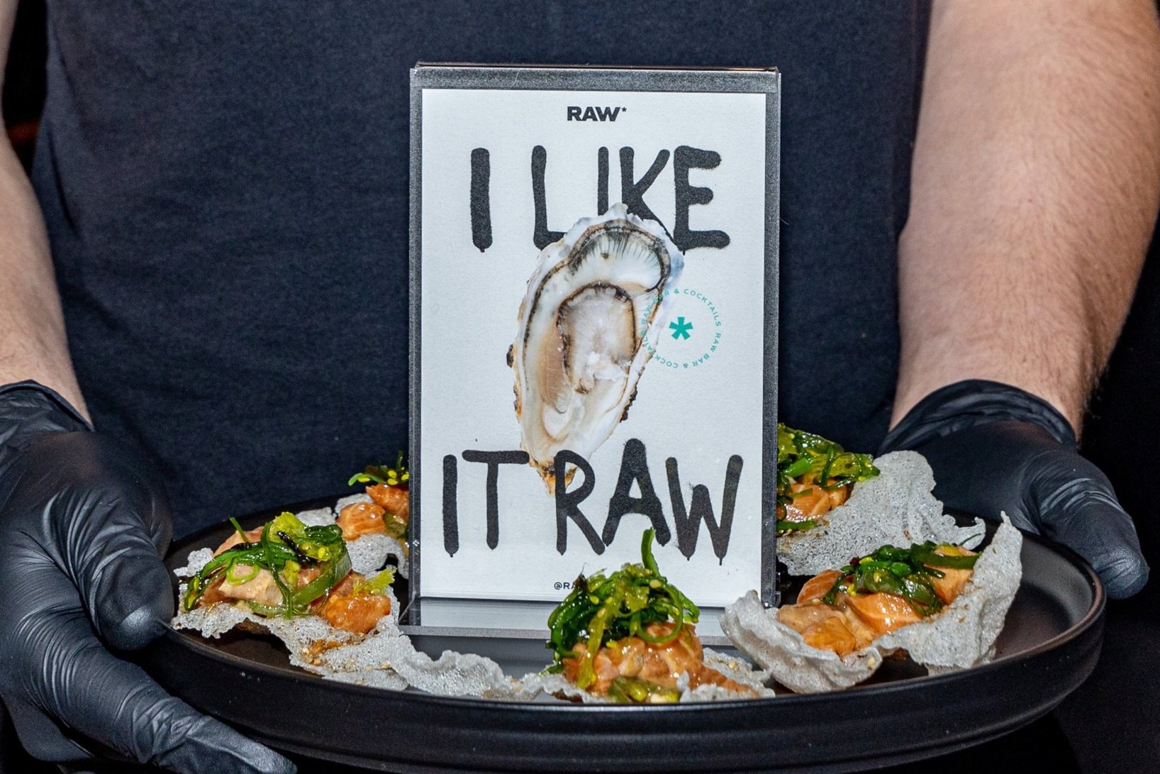 Stylized food presentation highlighting RAW's branding: a clean card with an oyster graphic and the phrase "I Like It Raw" takes center stage, accompanied by a visually appealing dish.