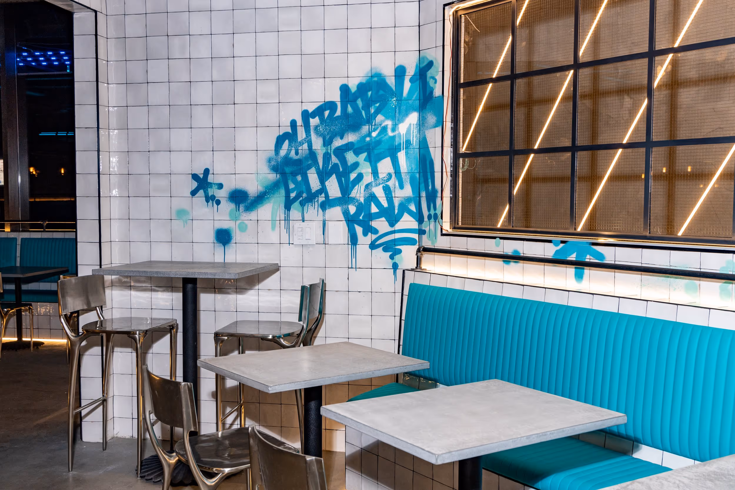 Interior view of RAW showcasing a textured white tiled wall with a vibrant blue graffiti mural, complemented by teal upholstered booth seating and minimalist metal chairs around gray tabletops.