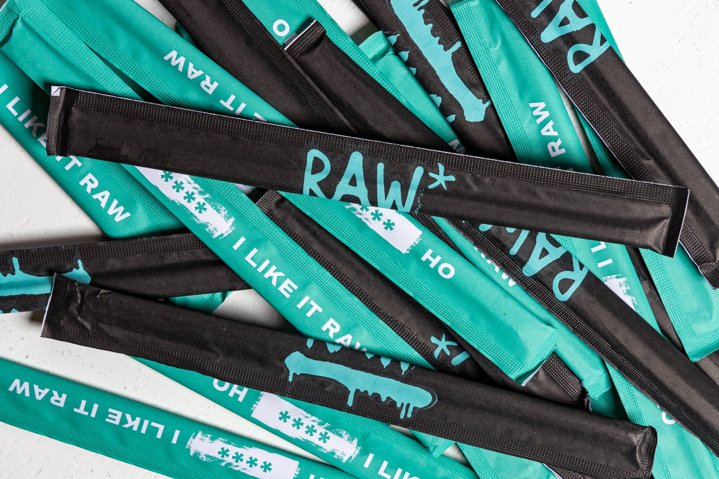 Detail-oriented view of RAW's packaging design, highlighting the contrasting teal and black color scheme, the prominent "RAW*" logo, and the playful tagline "I Like It Raw".