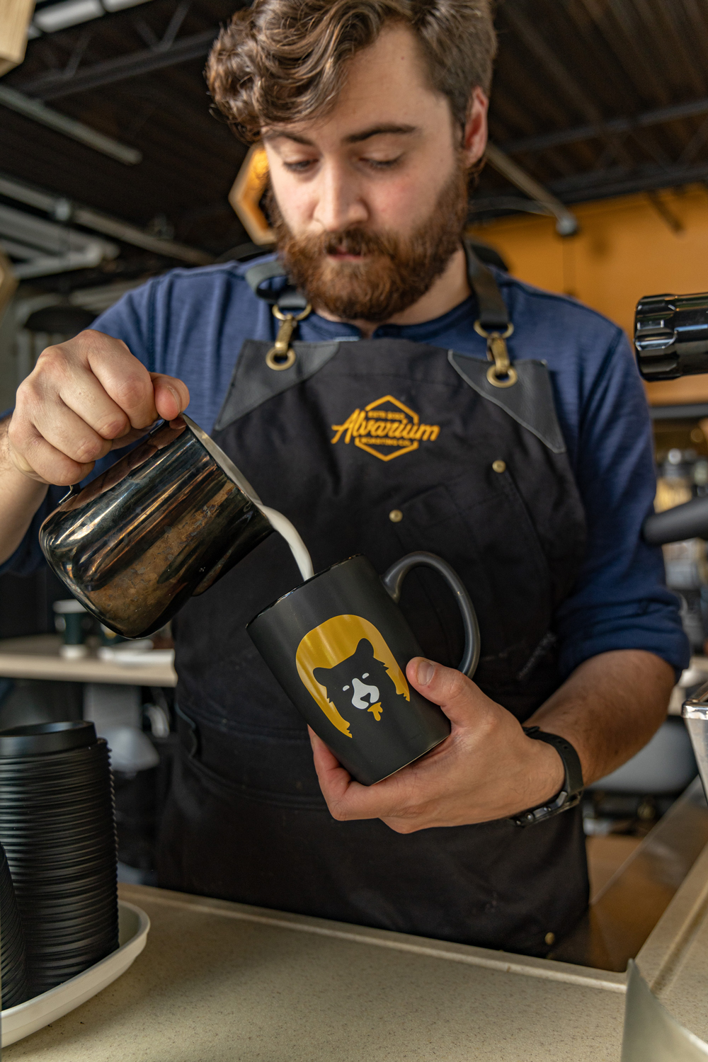 Photo of a beverage preparation, emphasizing the combination of the barista's action, the creamy milk being poured, and the Alvarium Roasting Co. branding on the mug.