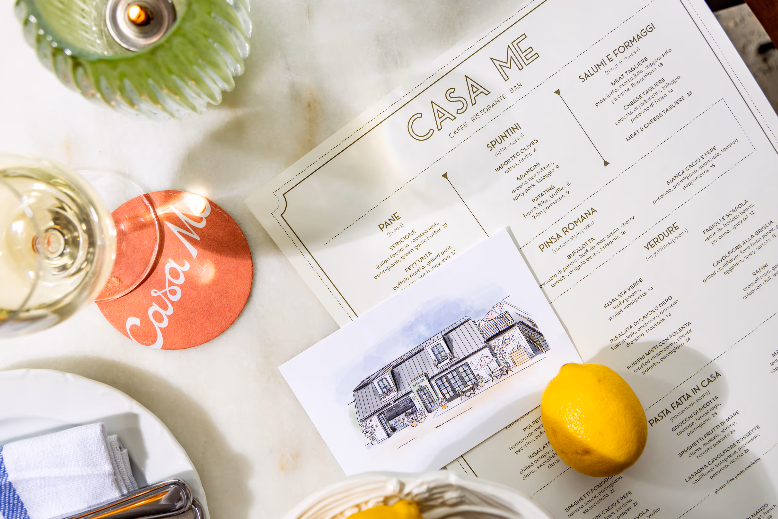 Flat lay of a Casa Me menu, coaster, illustrated card, and table setting elements, showcasing the restaurant's branding and dining experience.