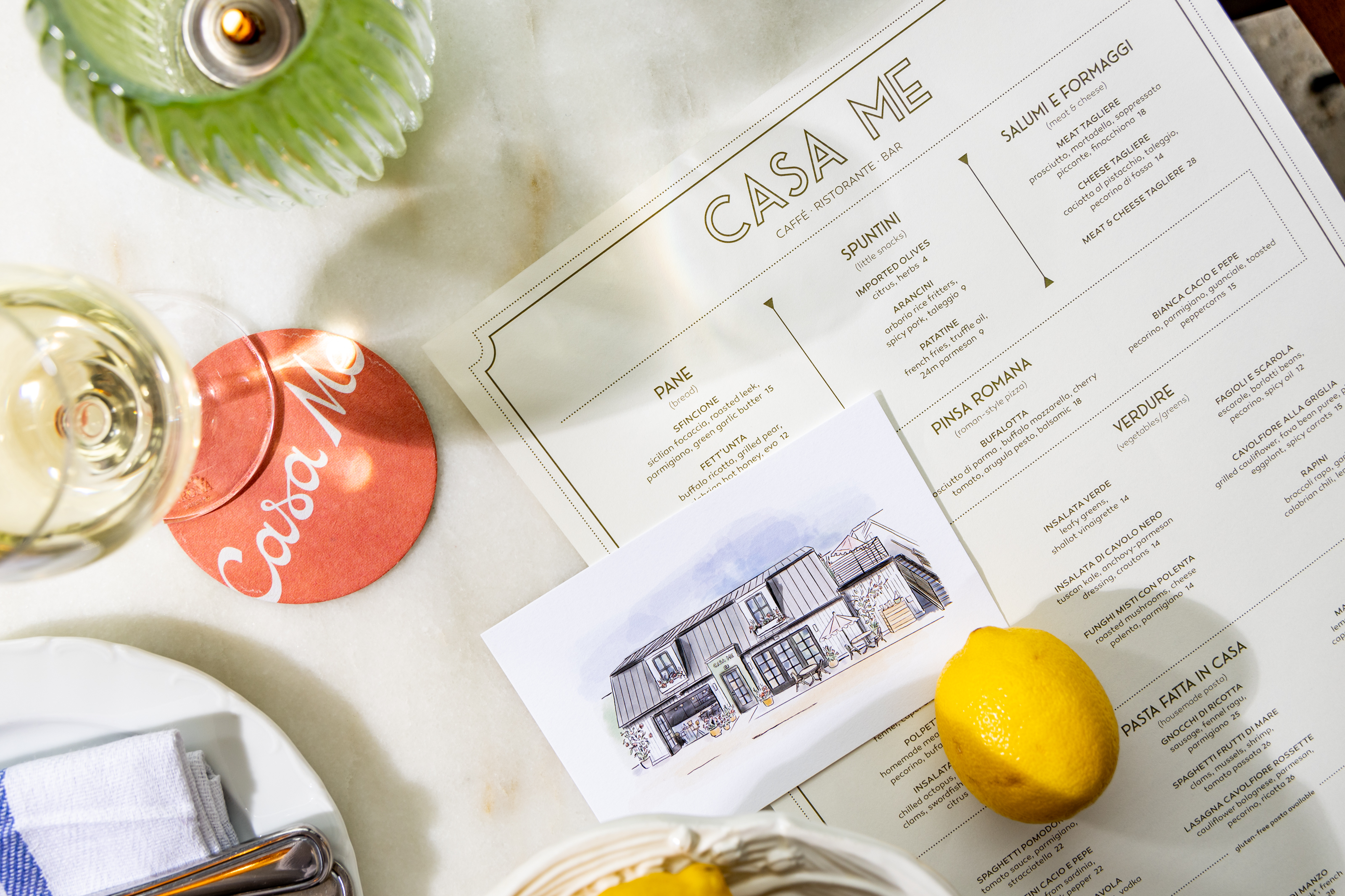 Flat lay of a Casa Me menu, coaster, illustrated card, and table setting elements, showcasing the restaurant's branding and dining experience.