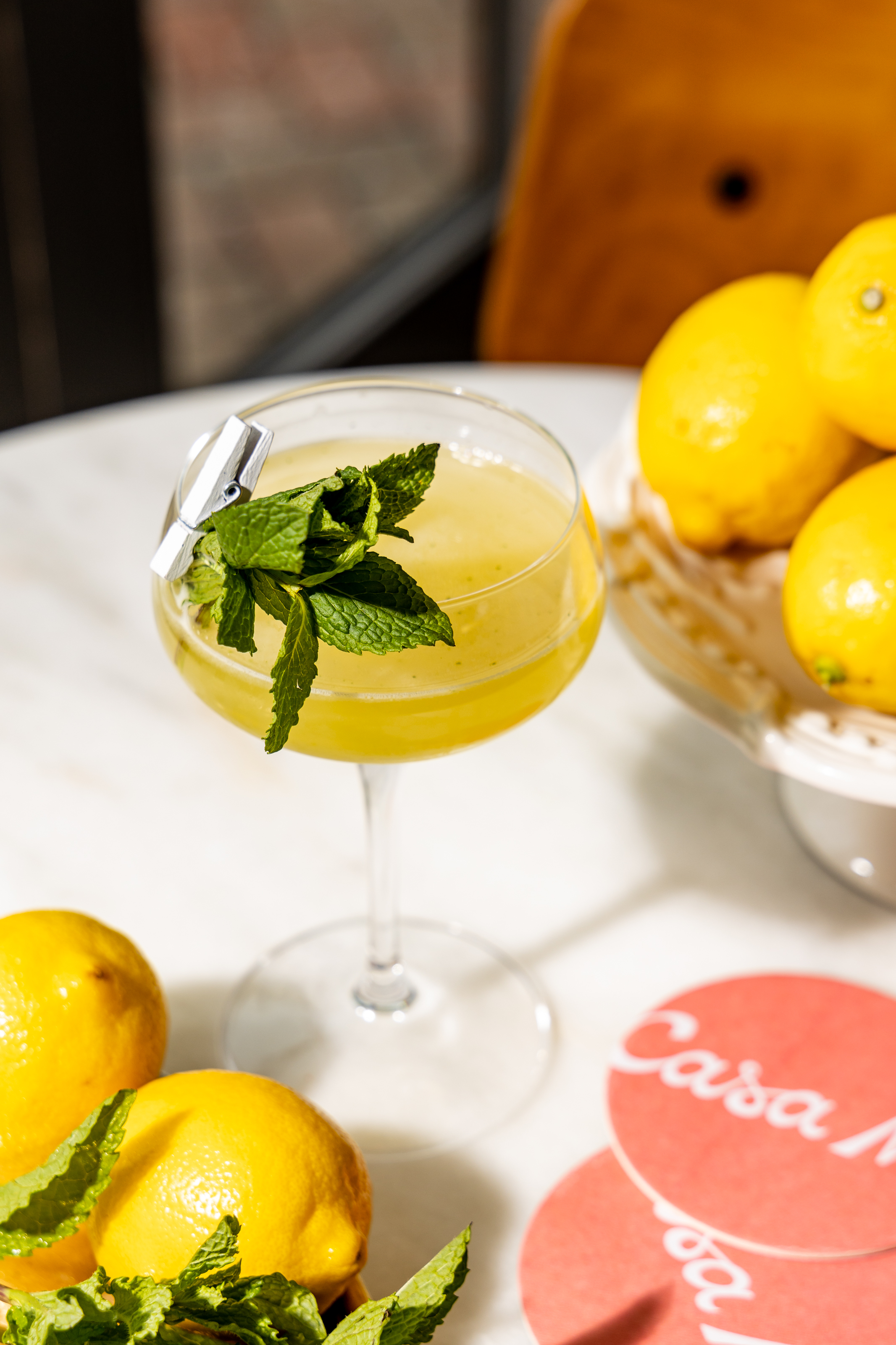 Close-up of a Casa Me branded cocktail in a coupe glass, garnished with mint and a small clip, surrounded by fresh lemons and Casa Me coasters, showcasing the drink's presentation and branding.