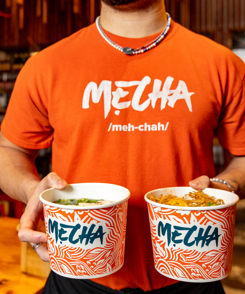Close-up of a Mecha Noodle Bar employee wearing an orange branded t-shirt and a necklace, holding two takeout containers featuring the restaurant's logo and stylized wave pattern.
