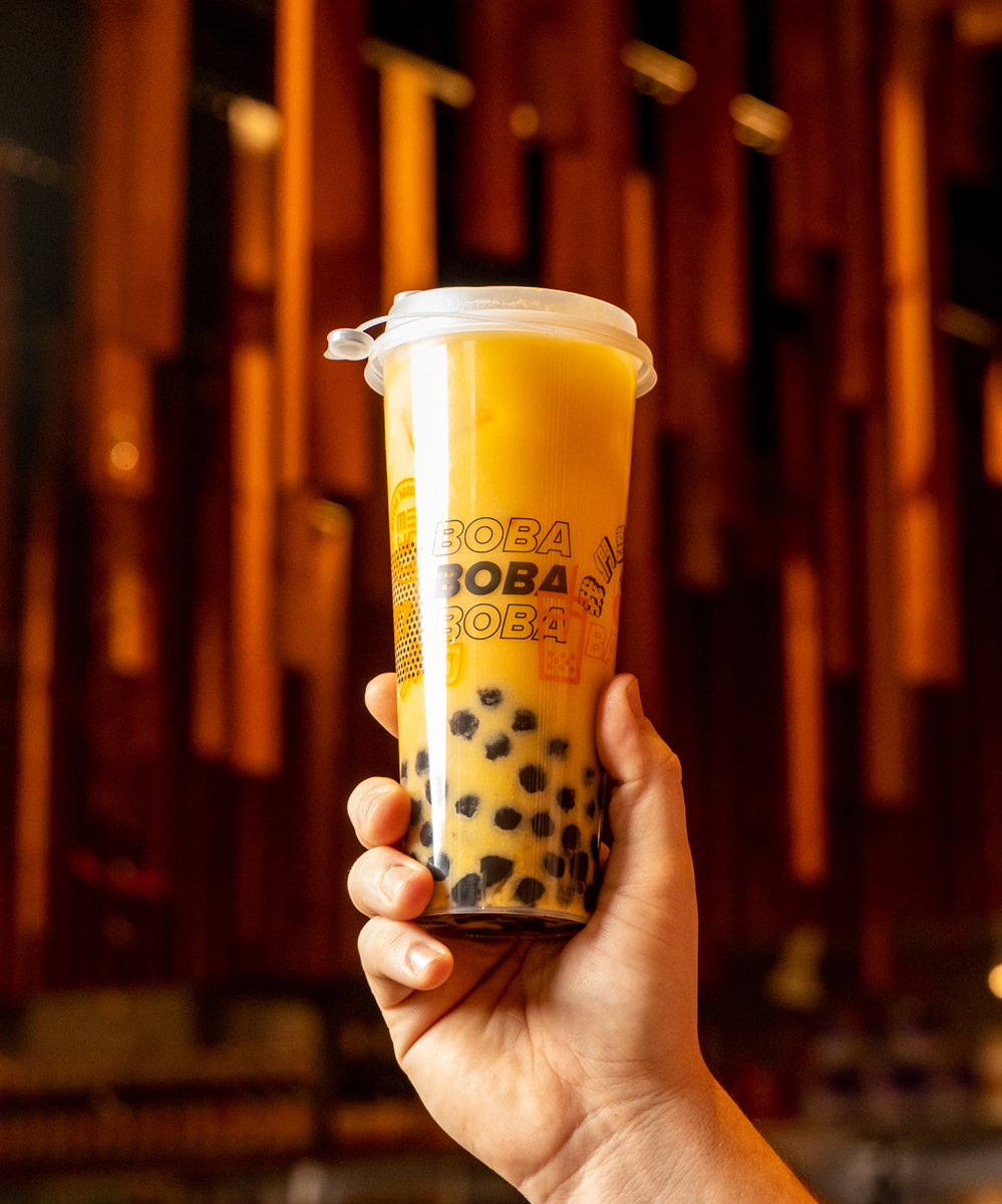 Close-up of a hand holding a clear plastic cup filled with a mango-flavored boba drink, showcasing the layered texture of the beverage with tapioca pearls at the bottom and the brand's repeating logo design on the side.