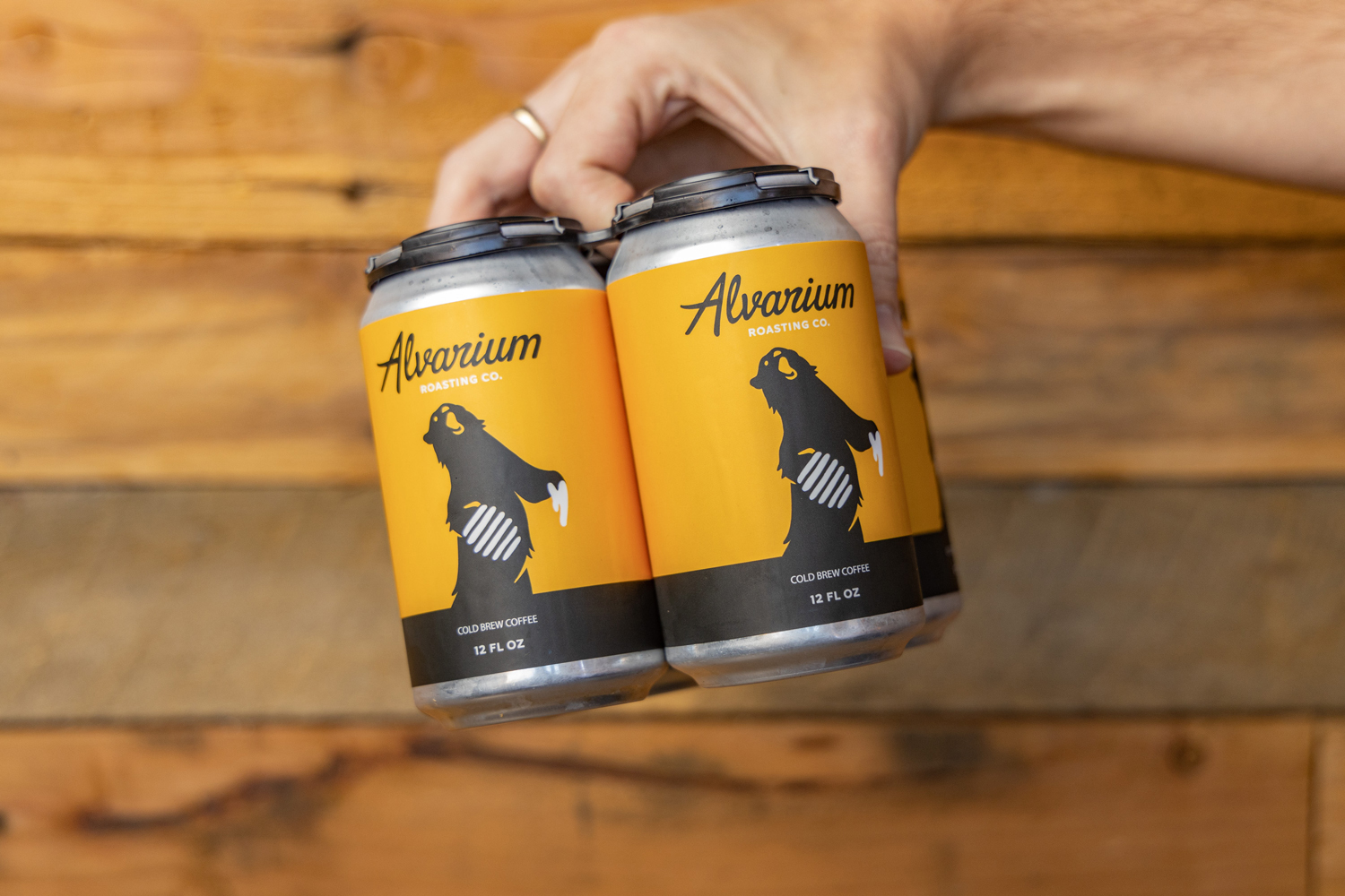 Image of a hand holding two yellow cans of Alvarium Roasting Co. cold brew coffee, showcasing the brand's bear logo and product information against a blurred wooden background.