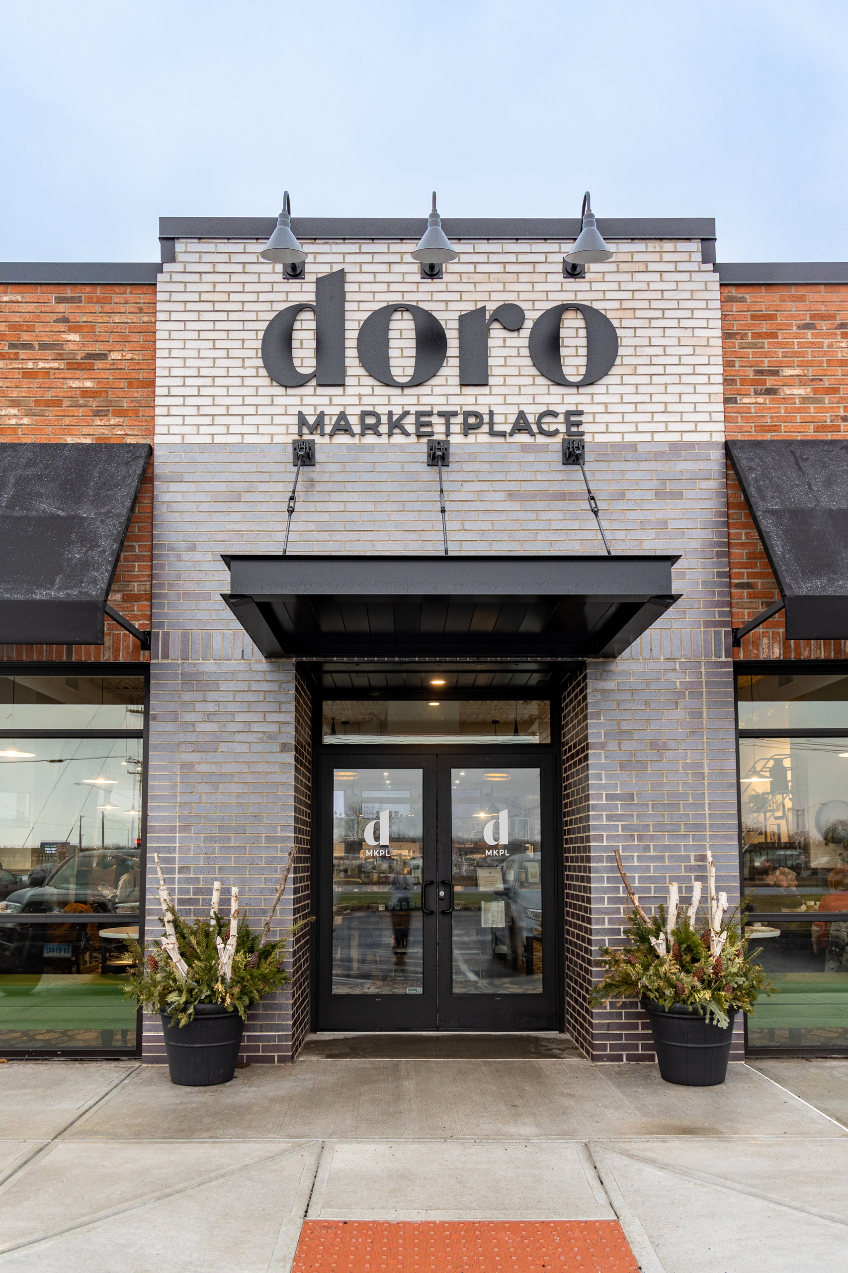A photo of the Doro Marketplace storefront. The building has a modern facade with a mix of brick and white siding. The "Doro Marketplace" sign is prominently displayed above the entrance, which is framed by black metal awnings.
