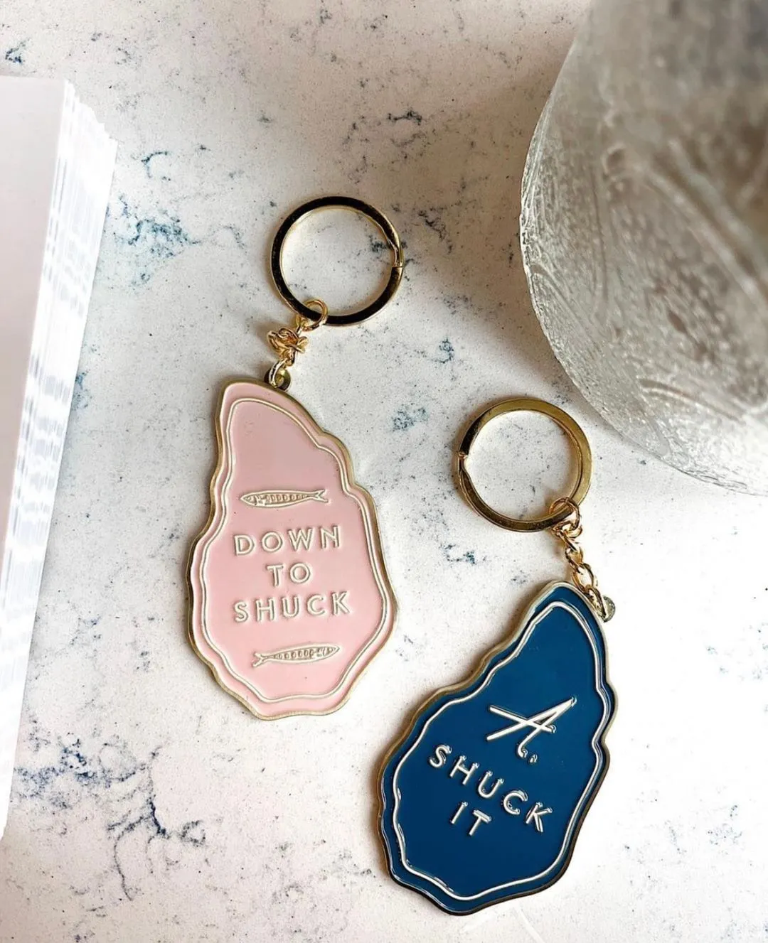 Image of two enamel keychains featuring the phrase "Down to Shuck" and a variation "A Shuck It," showcasing a playful design with contrasting colors and a nod to oyster shucking.