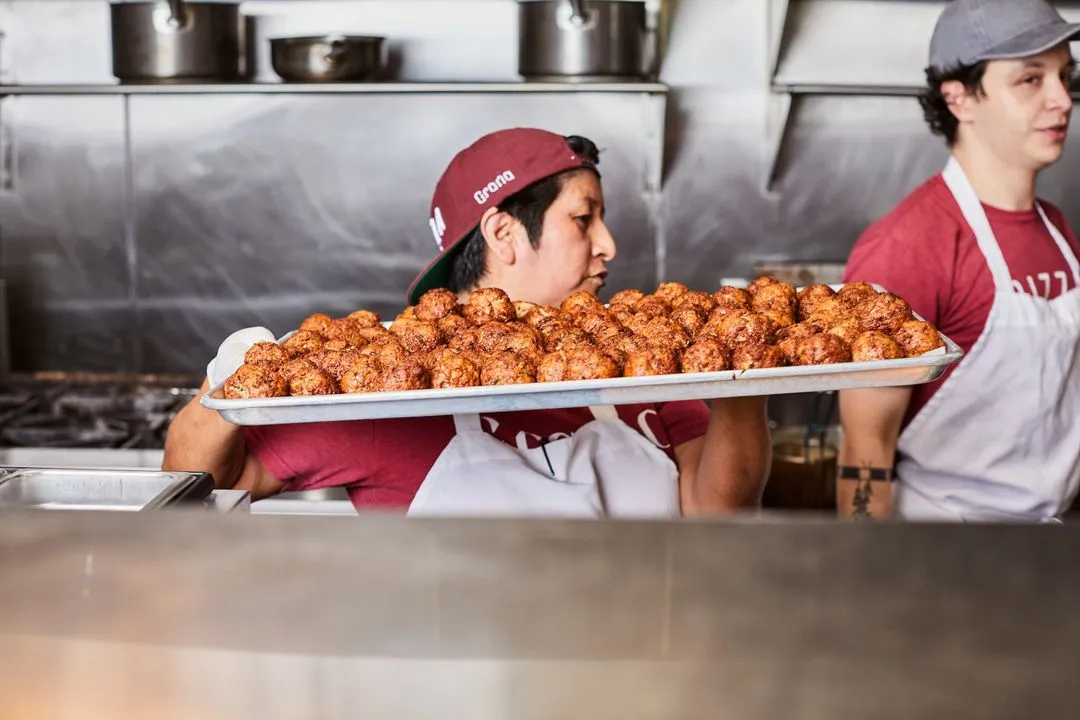 Photograph of a rectangular, metal serving tray generously filled with cooked meatballs being held by a person wearing a red shirt and white apron.